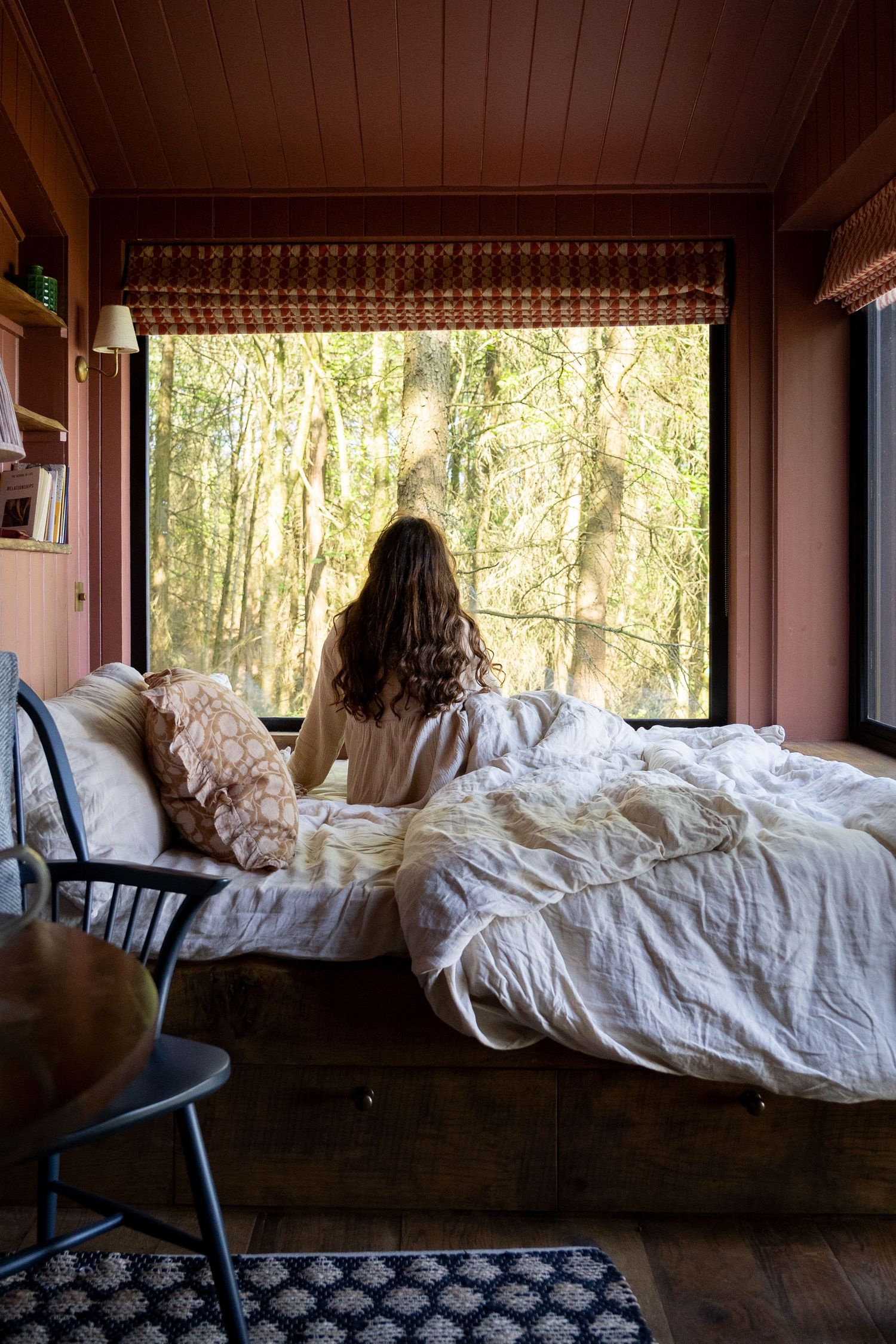 Woman sitting on a bed with white linen looking out a large window into a sunlit forest in a cozy cabin.