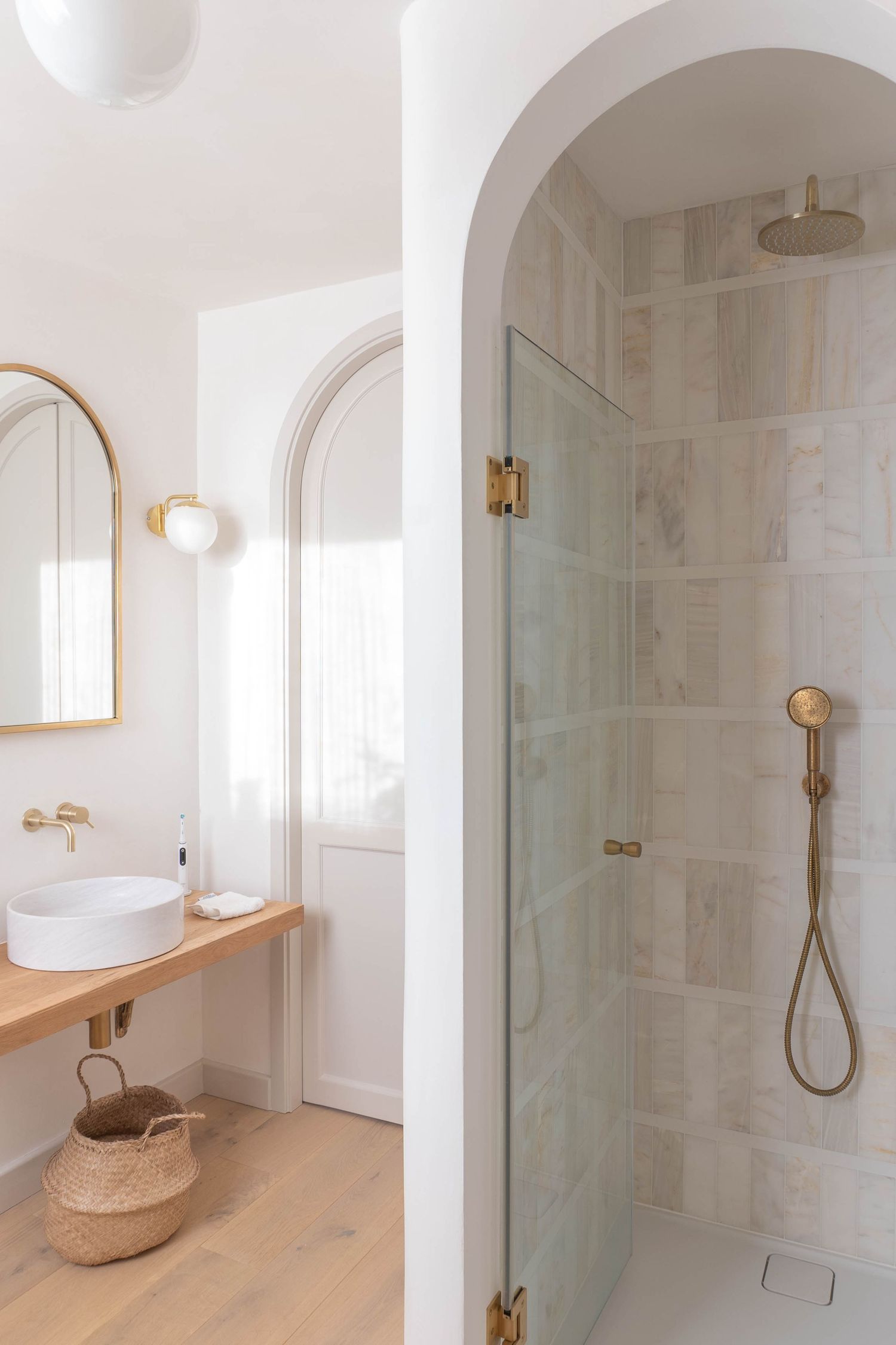 Modern white bathroom with arched shower, brass fixtures, wood vanity, vessel sink, and gold-framed mirror.