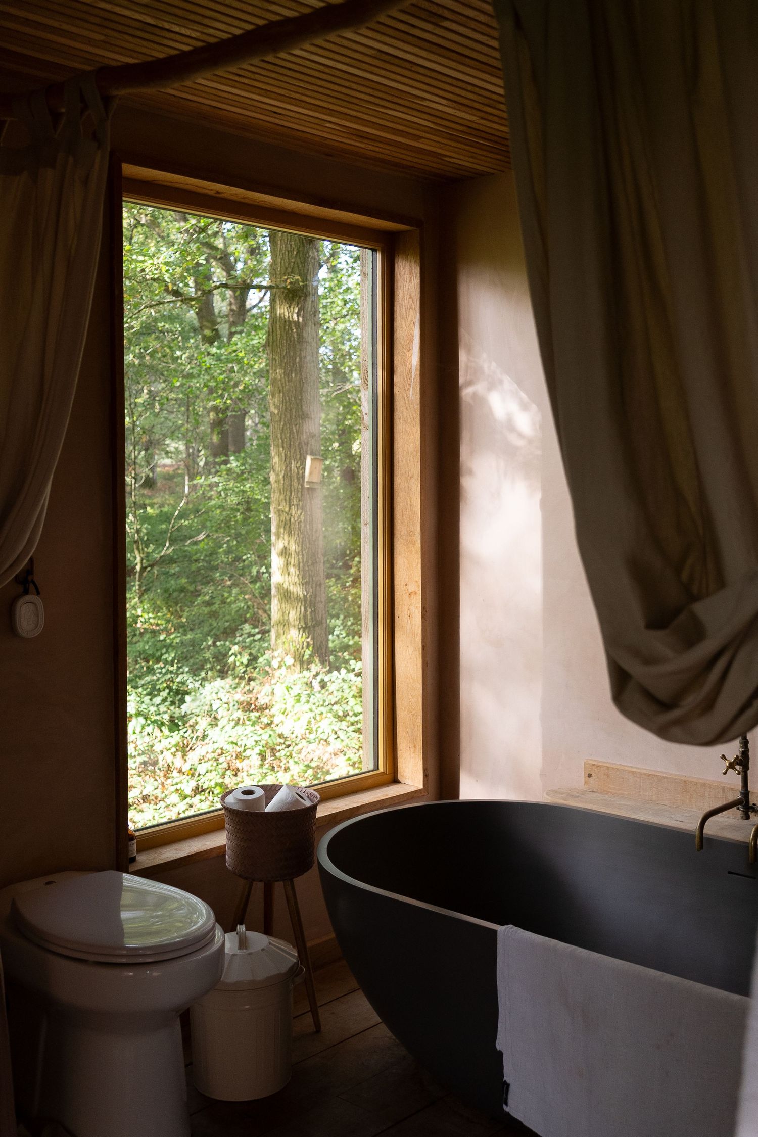 Rustic bathroom with dark freestanding bathtub, linen curtains, and large wooden-framed window overlooking lush green forest.