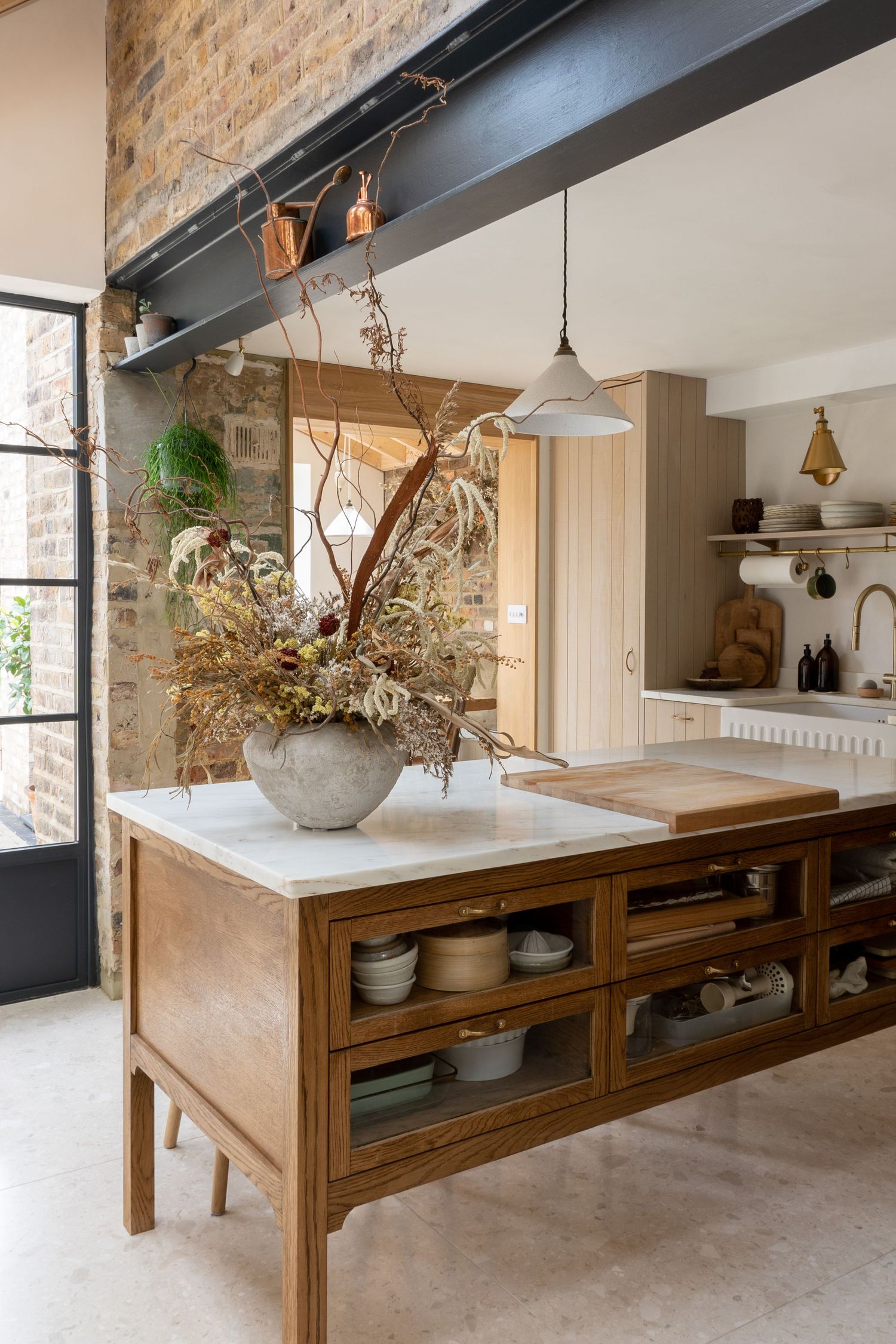 Rustic kitchen with exposed brick wall, wooden island with glass cabinets, and large dried botanical arrangement.
