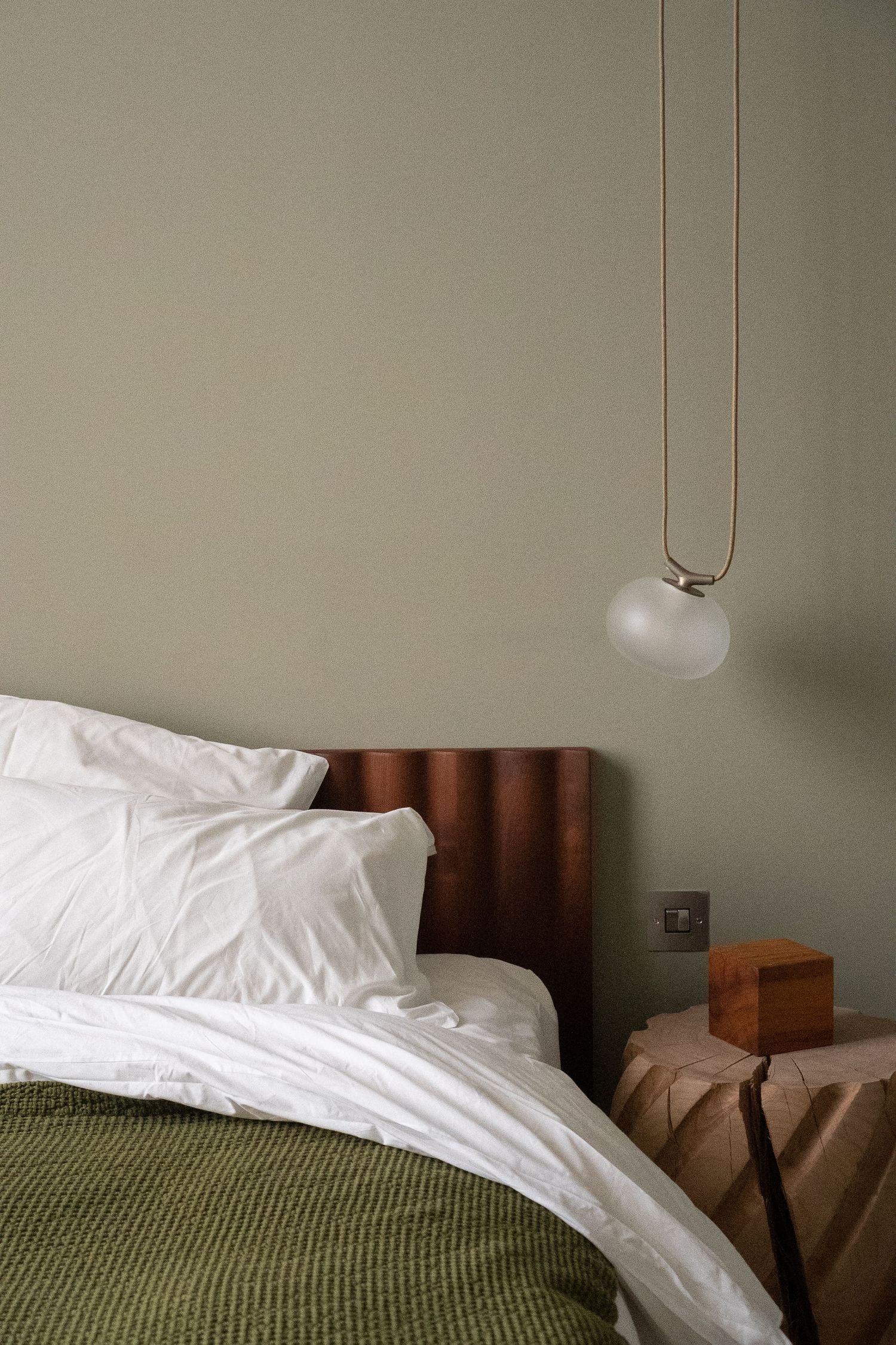Minimalist bedroom with sage green walls, white bedding, walnut headboard, and frosted glass pendant light.