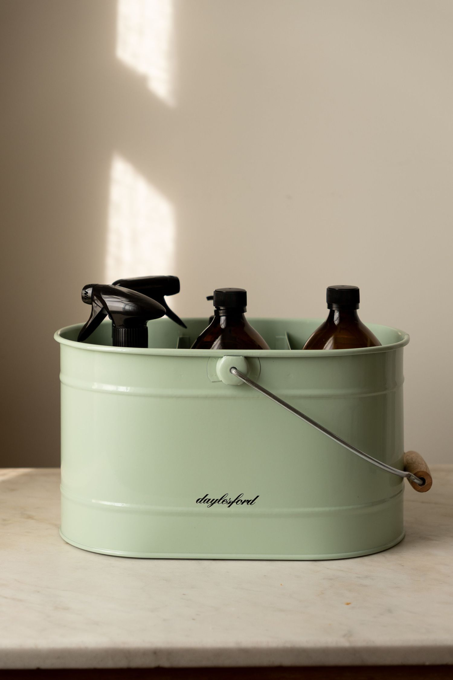 Daylesford branded sage green metal bucket organizer holding amber glass cleaning spray bottles on marble surface.