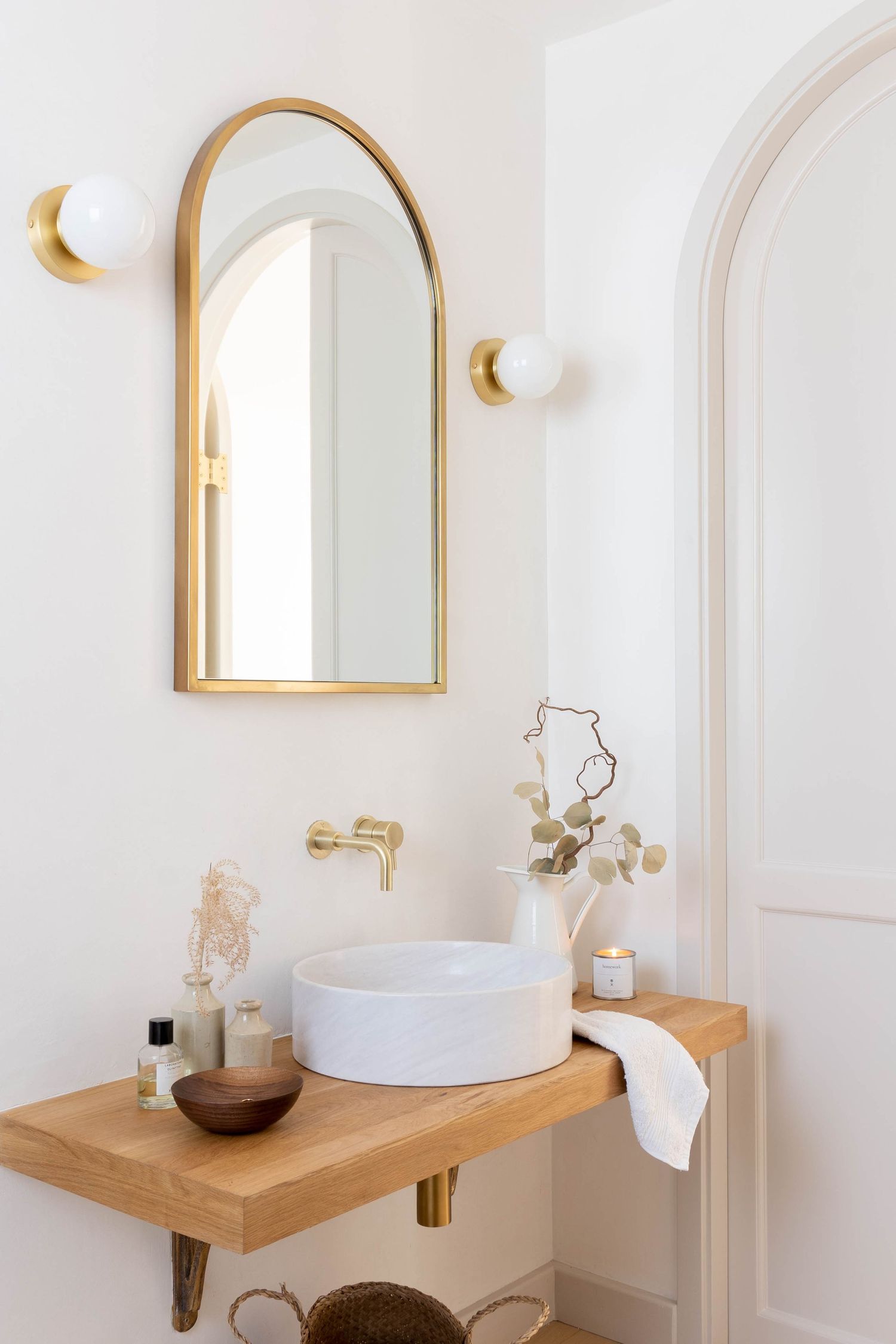 Elegant bathroom vanity with arched gold mirror, brass wall faucet, vessel sink on oak shelf, and globe sconces.