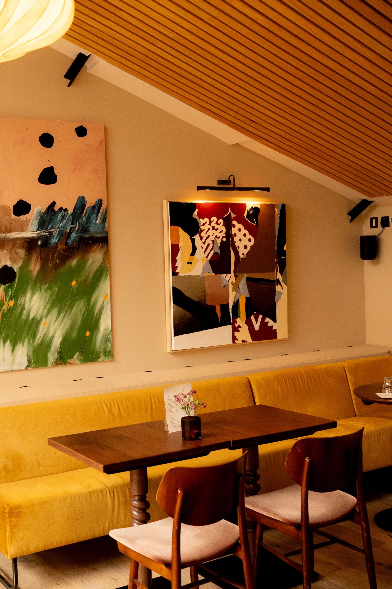 Modern restaurant dining area with mustard yellow velvet banquette, wooden tables, and colorful abstract wall art.