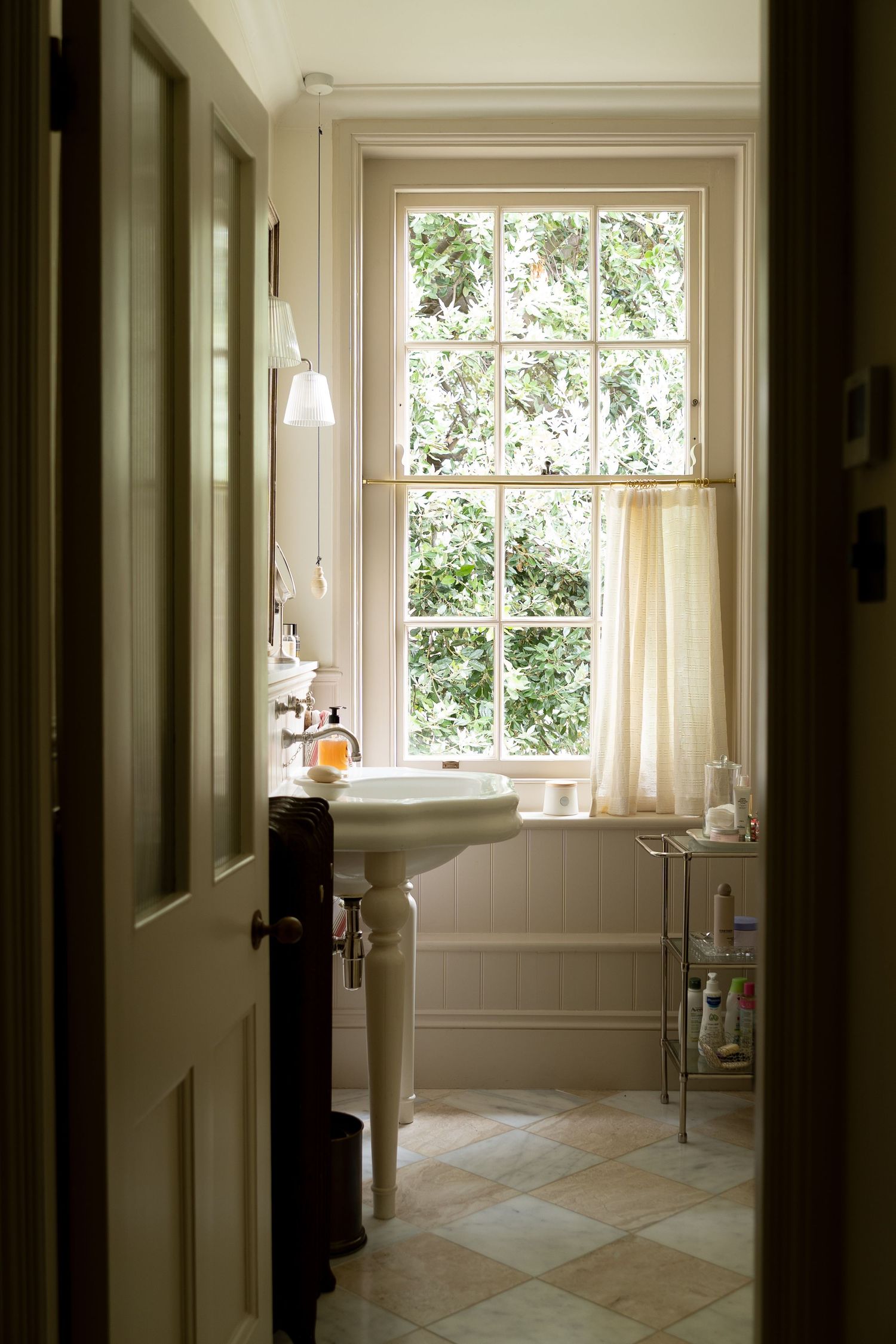 Classic white pedestal sink bathroom with large sash window overlooking green trees and checkered tile floor.