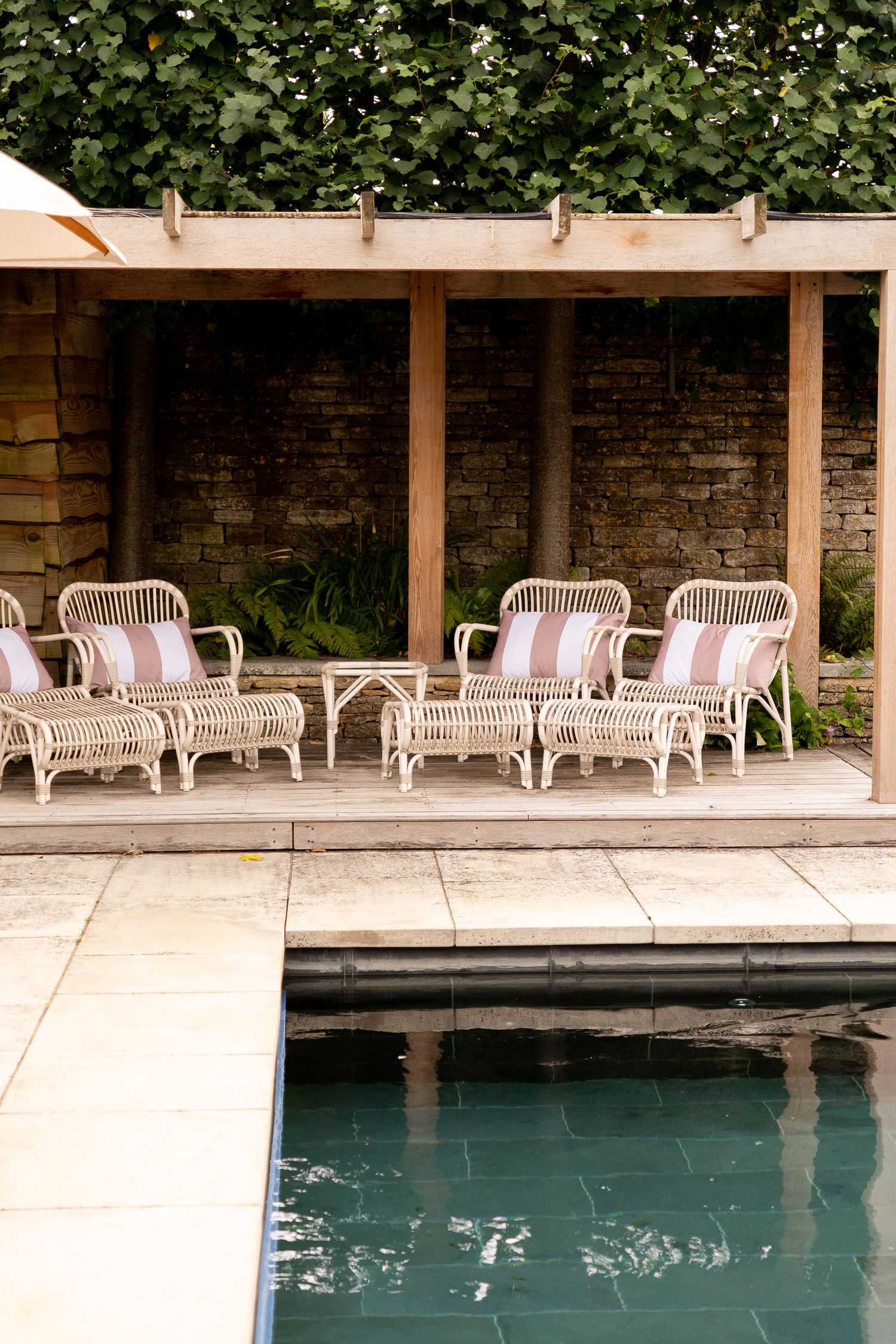 Poolside cabana with white rattan chairs, pink striped cushions, wooden pergola, and stone wall surrounded by greenery.
