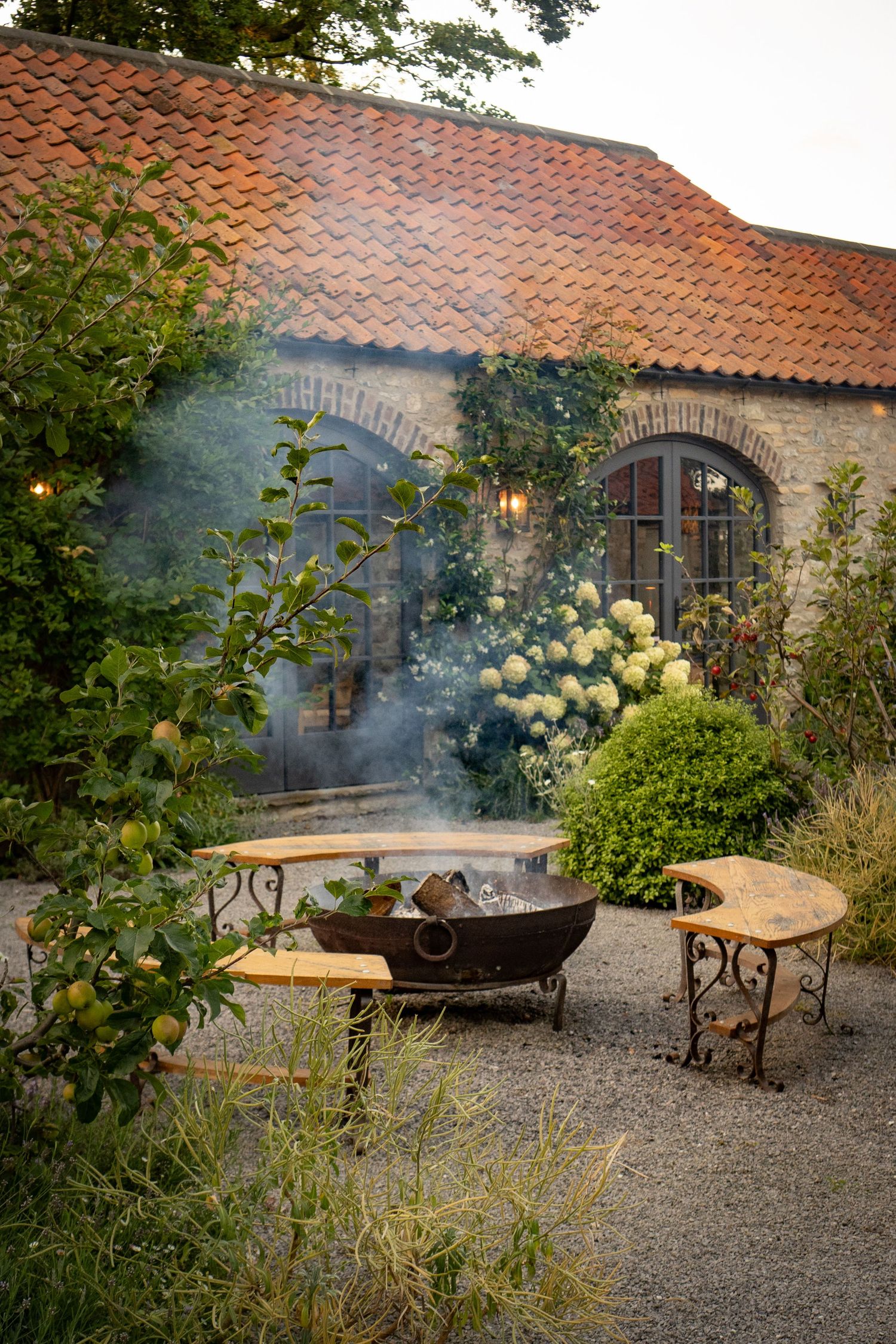 Rustic garden courtyard with smoking fire pit beside a stone building with terracotta roof.