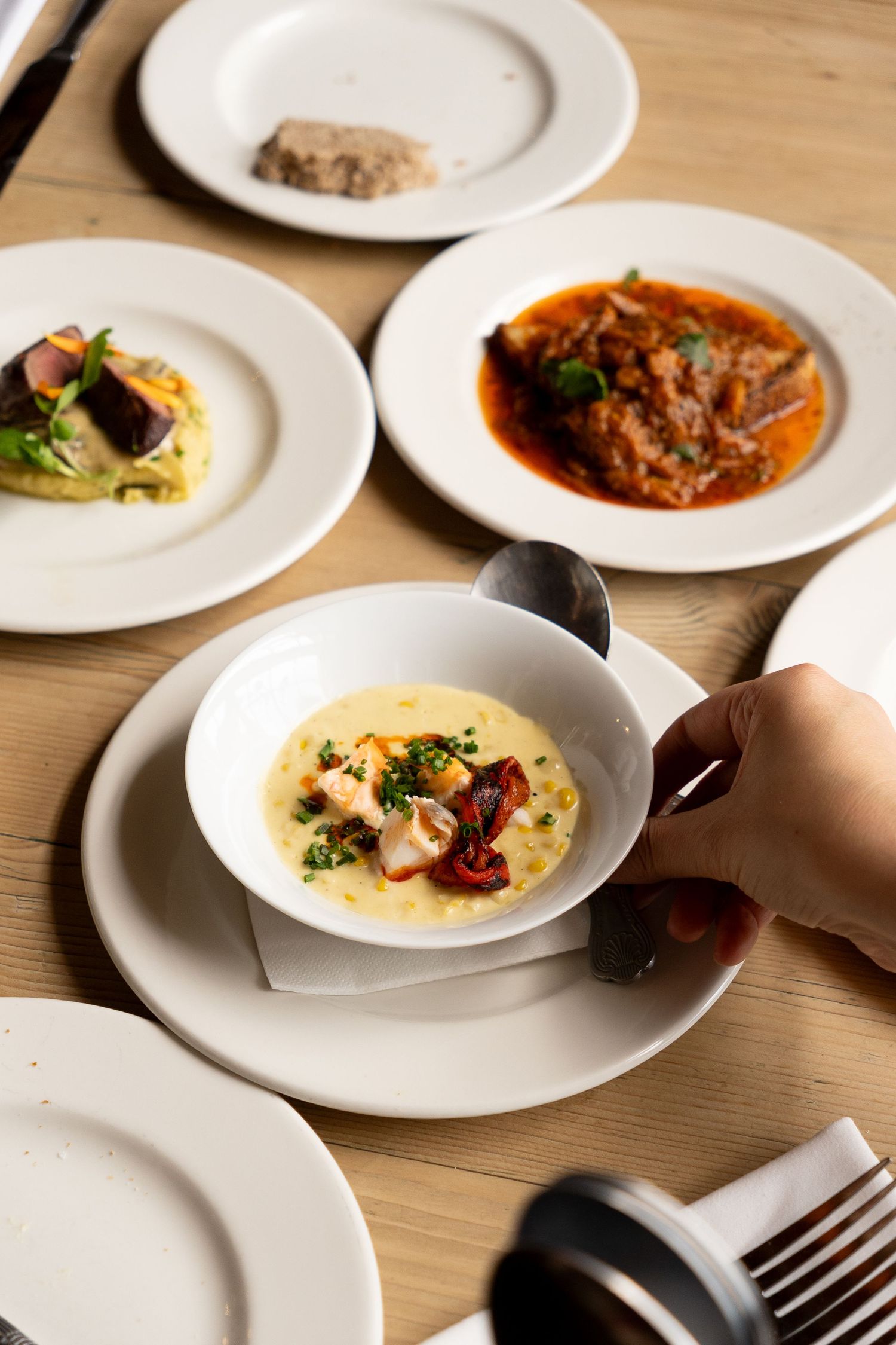 Hand holding white bowl of creamy seafood chowder with octopus, surrounded by various gourmet dishes on wooden table.