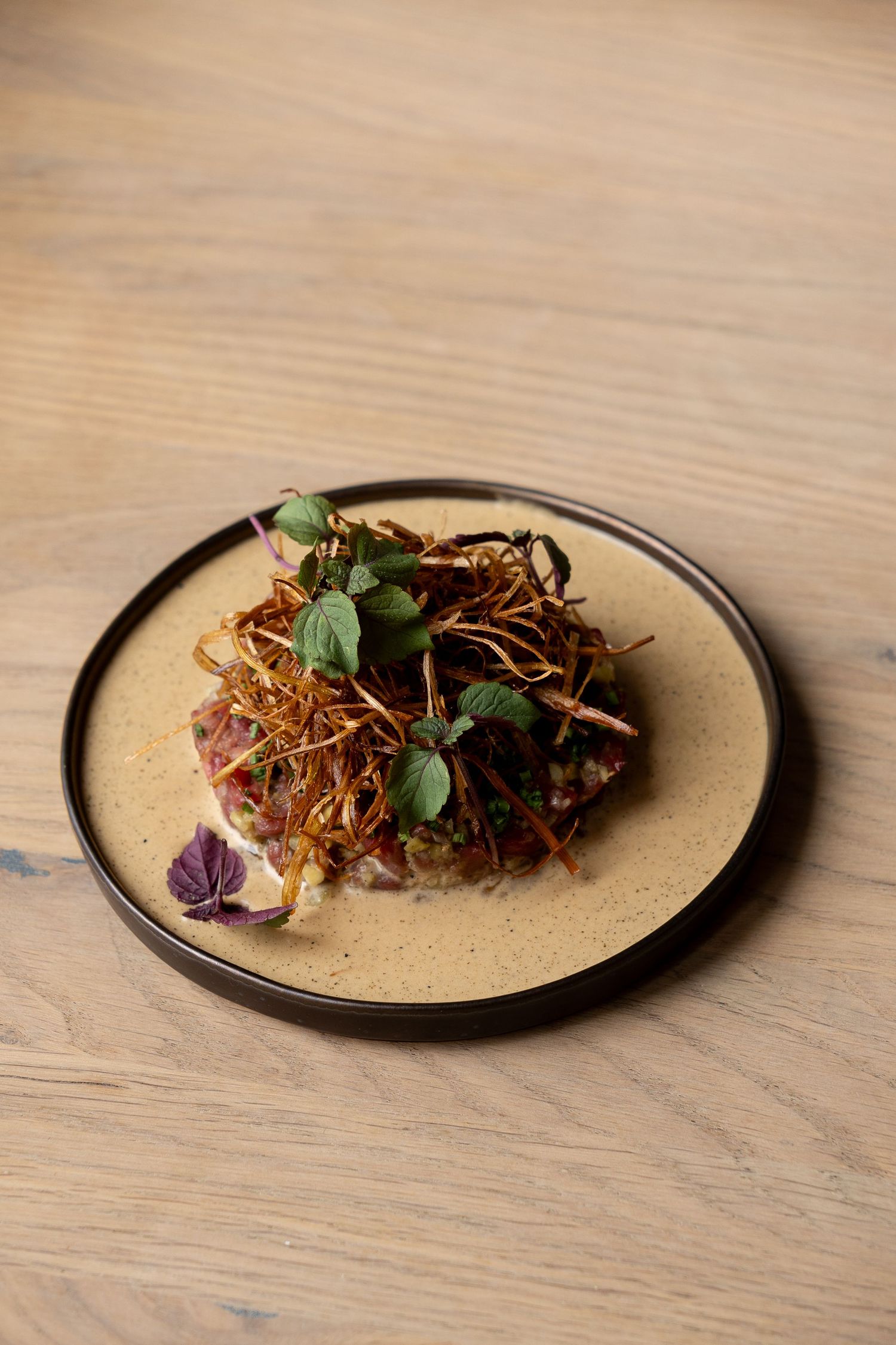 Elegant fine dining dish with crispy julienne strips, fresh microgreens, and tartare on a ceramic plate atop wooden table.