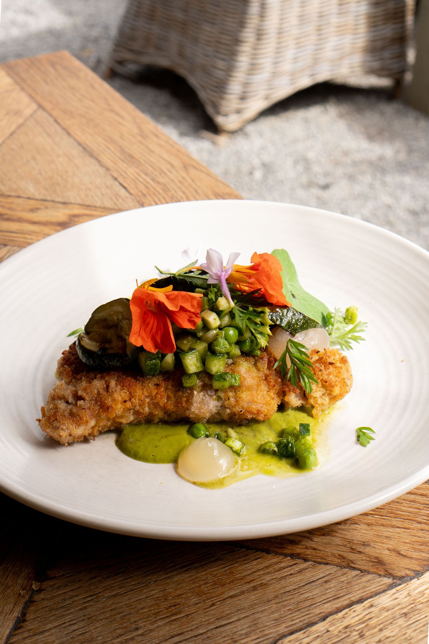 A crispy breaded fish fillet with green pea puree, vegetables, and vibrant edible flowers on a white ceramic plate.