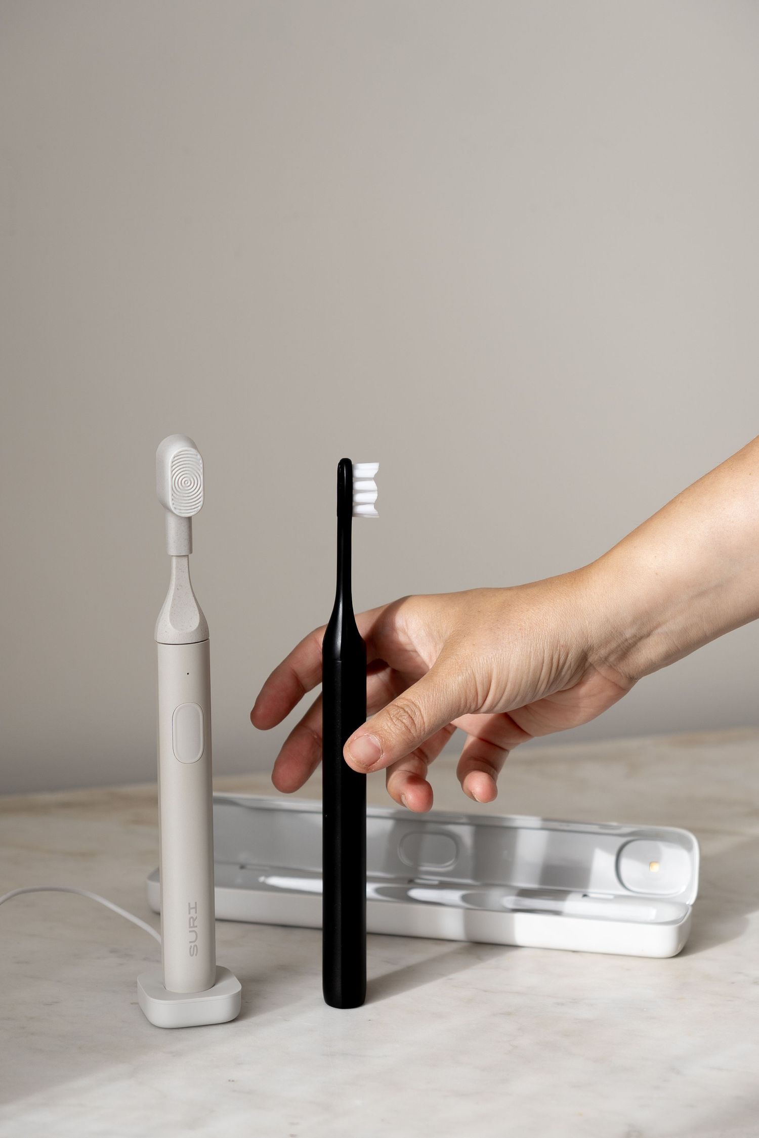 Hand reaching for a black electric toothbrush beside a white SURI toothbrush and travel case.