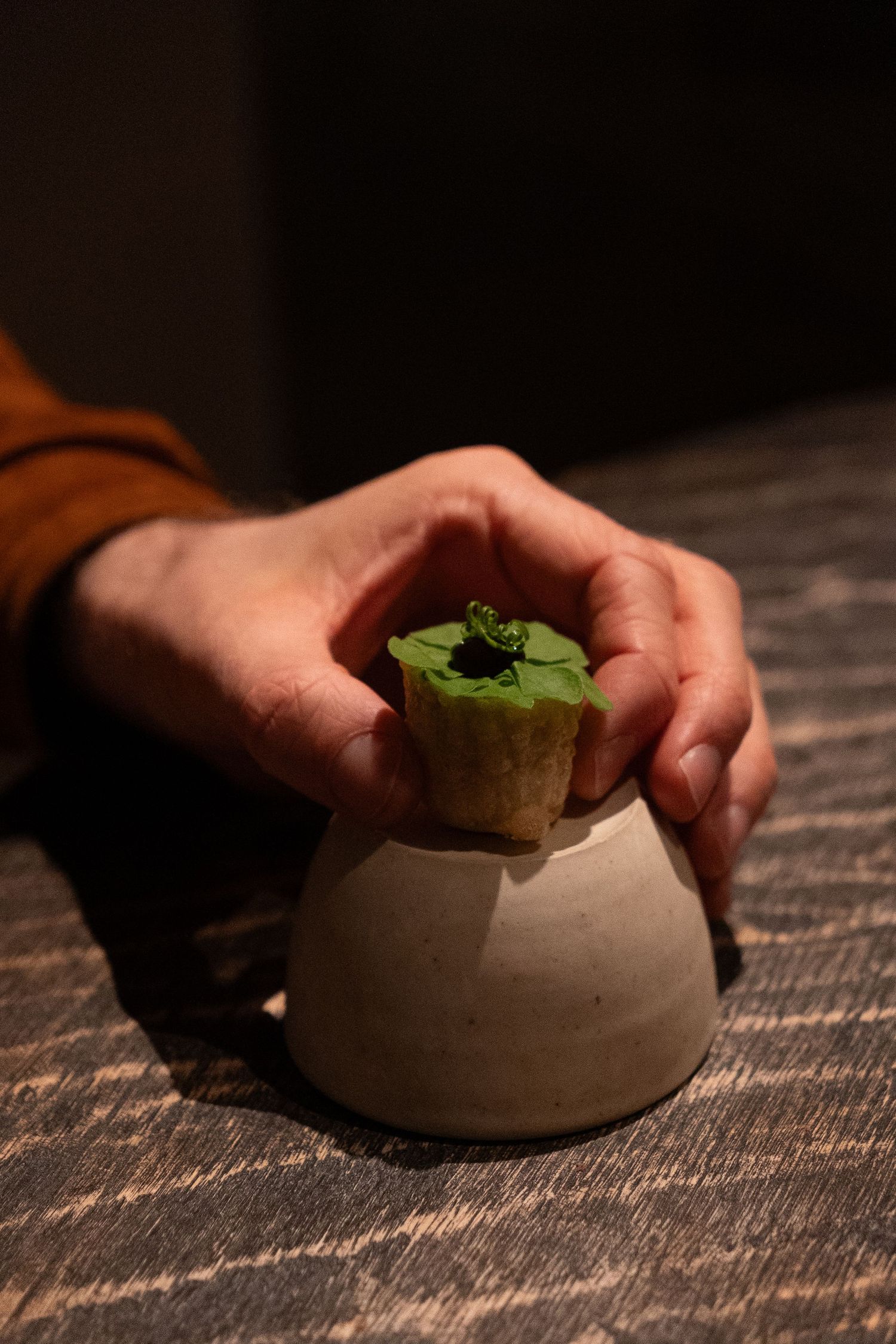 Hand placing an elegant green amuse-bouche on a ceramic pedestal at a fine dining restaurant.