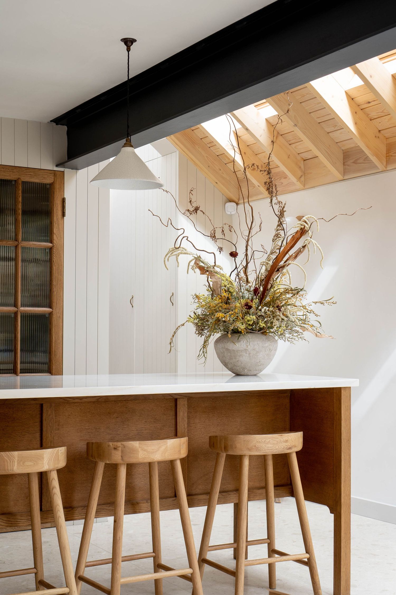 Modern kitchen island with wooden bar stools, dramatic dried floral centerpiece, and exposed timber ceiling beams.