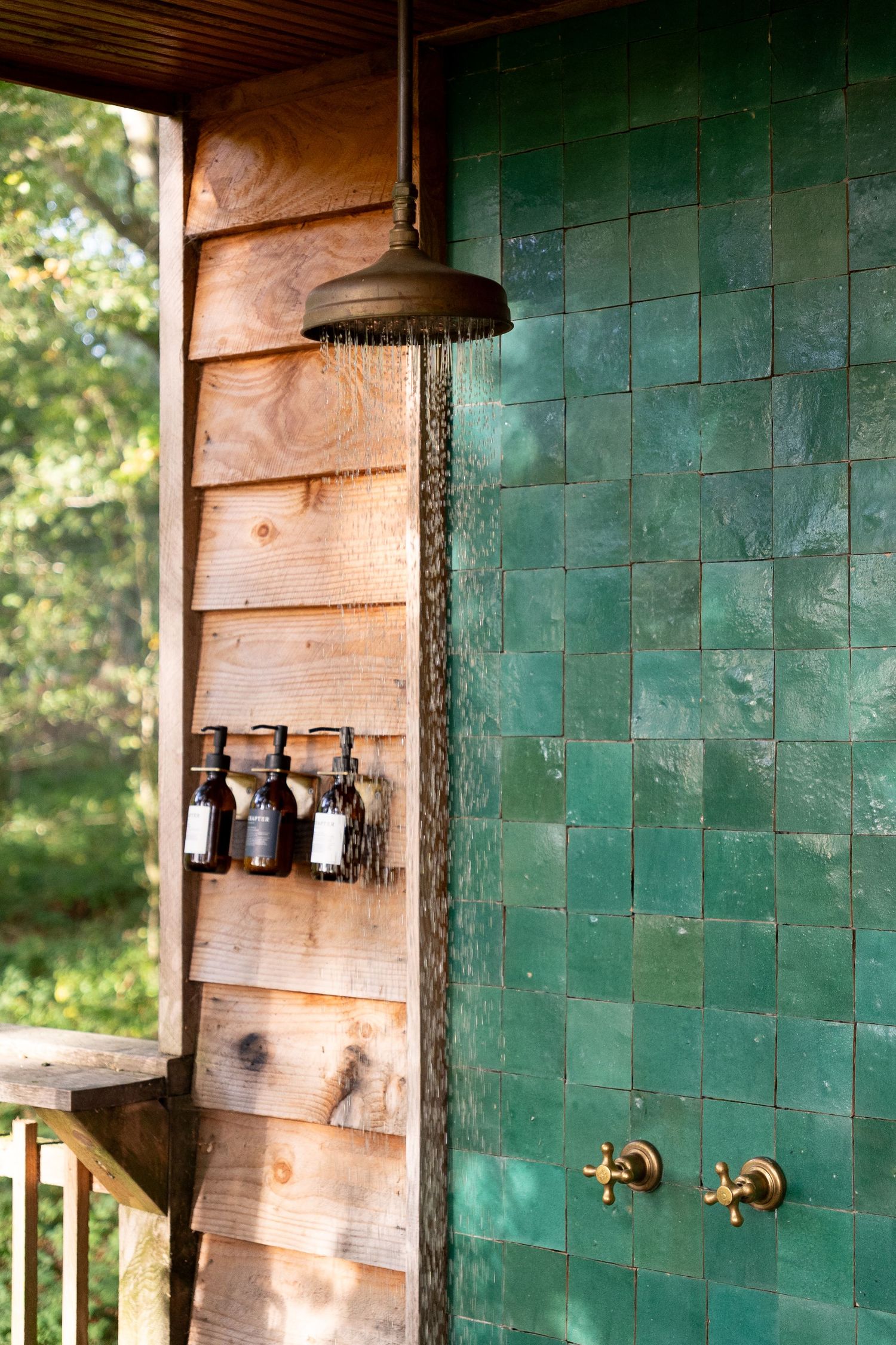 Outdoor shower with running water, green zellige tiles, brass fixtures, and wooden cabin wall with hanging toiletry bottles.