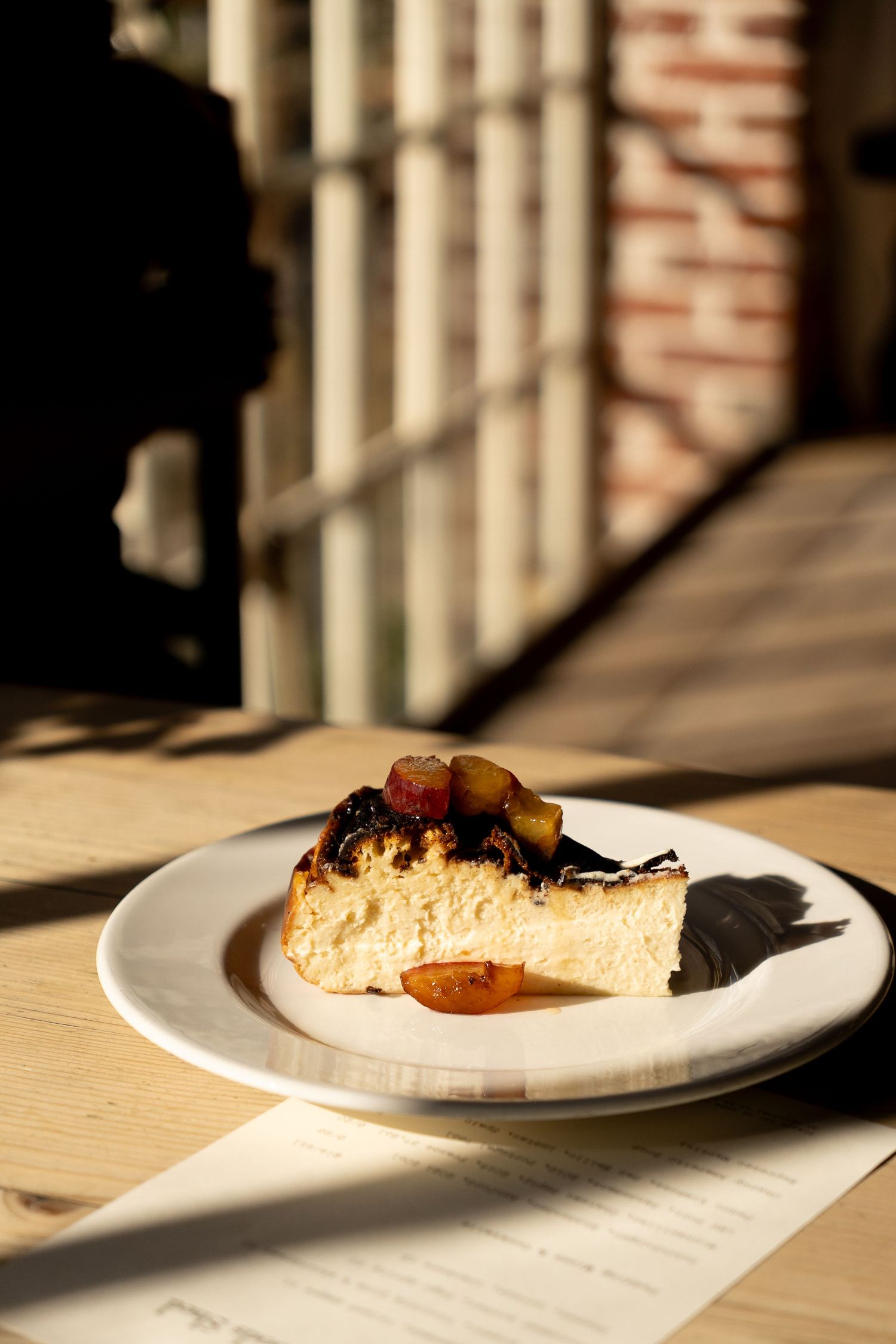 Slice of burnt Basque cheesecake with caramelized fruit topping on white plate in dramatic sunlight and shadows.