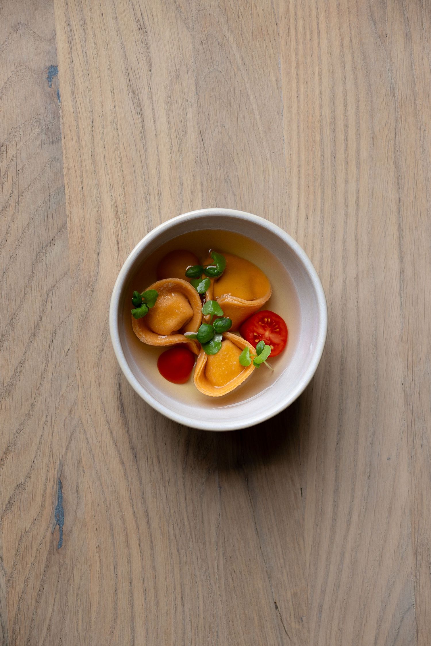 A white bowl of tortellini soup with cherry tomatoes and microgreens on a wooden surface.