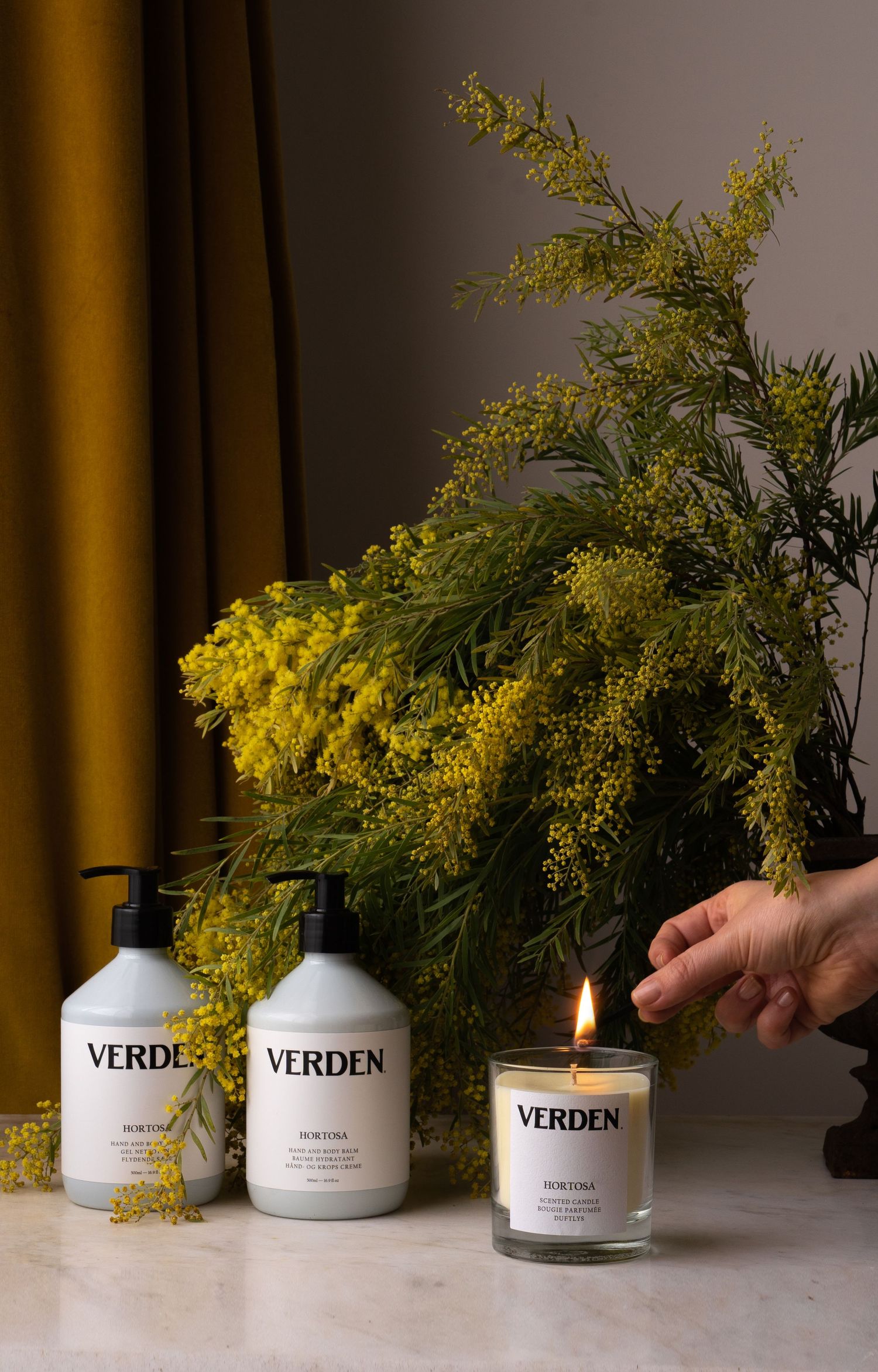 Verden Hortosa skincare collection with two white bottles and lit candle beside fresh yellow mimosa branches.