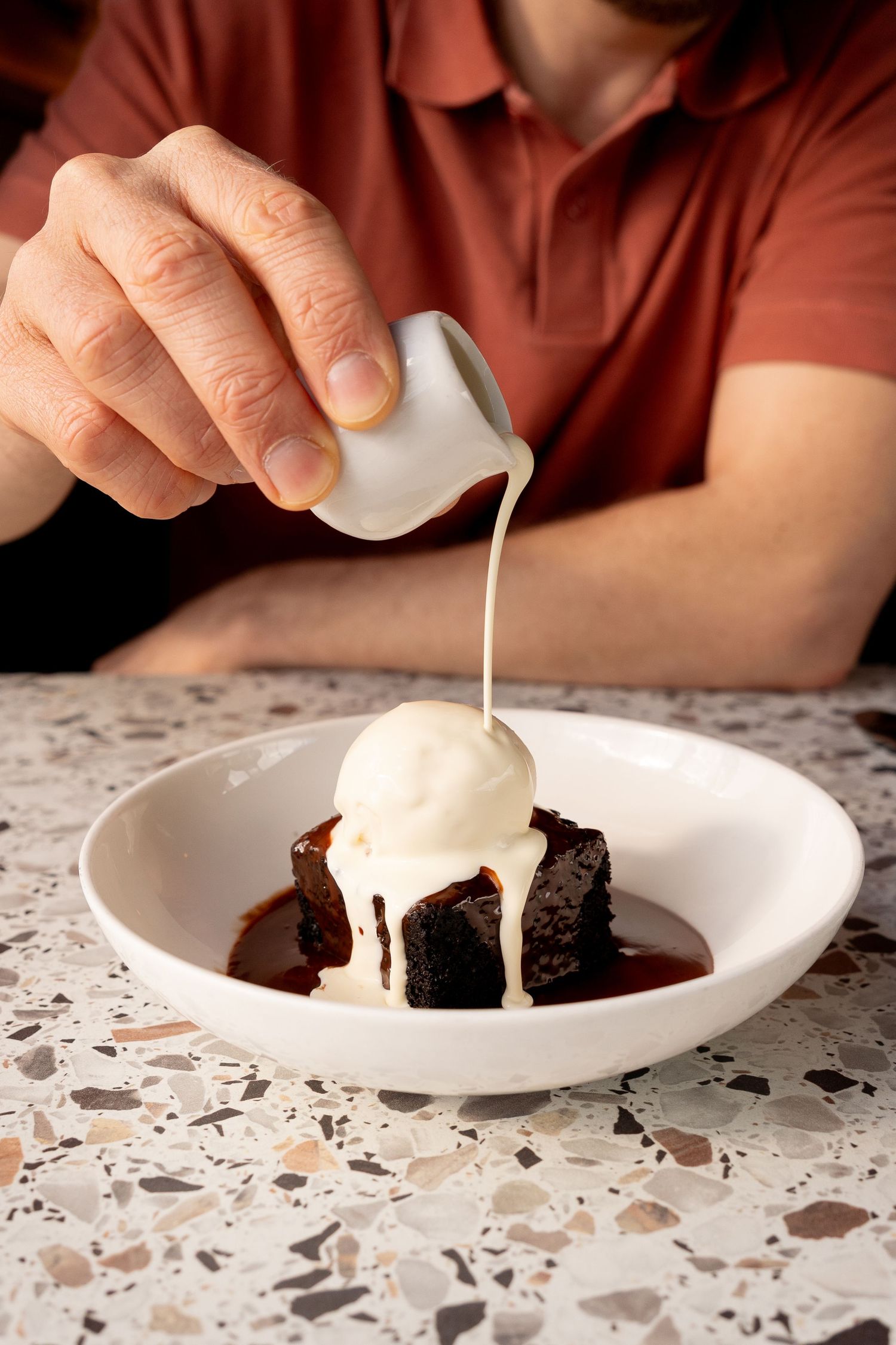 Person pouring cream over sticky date pudding with vanilla ice cream in a white bowl on terrazzo table.