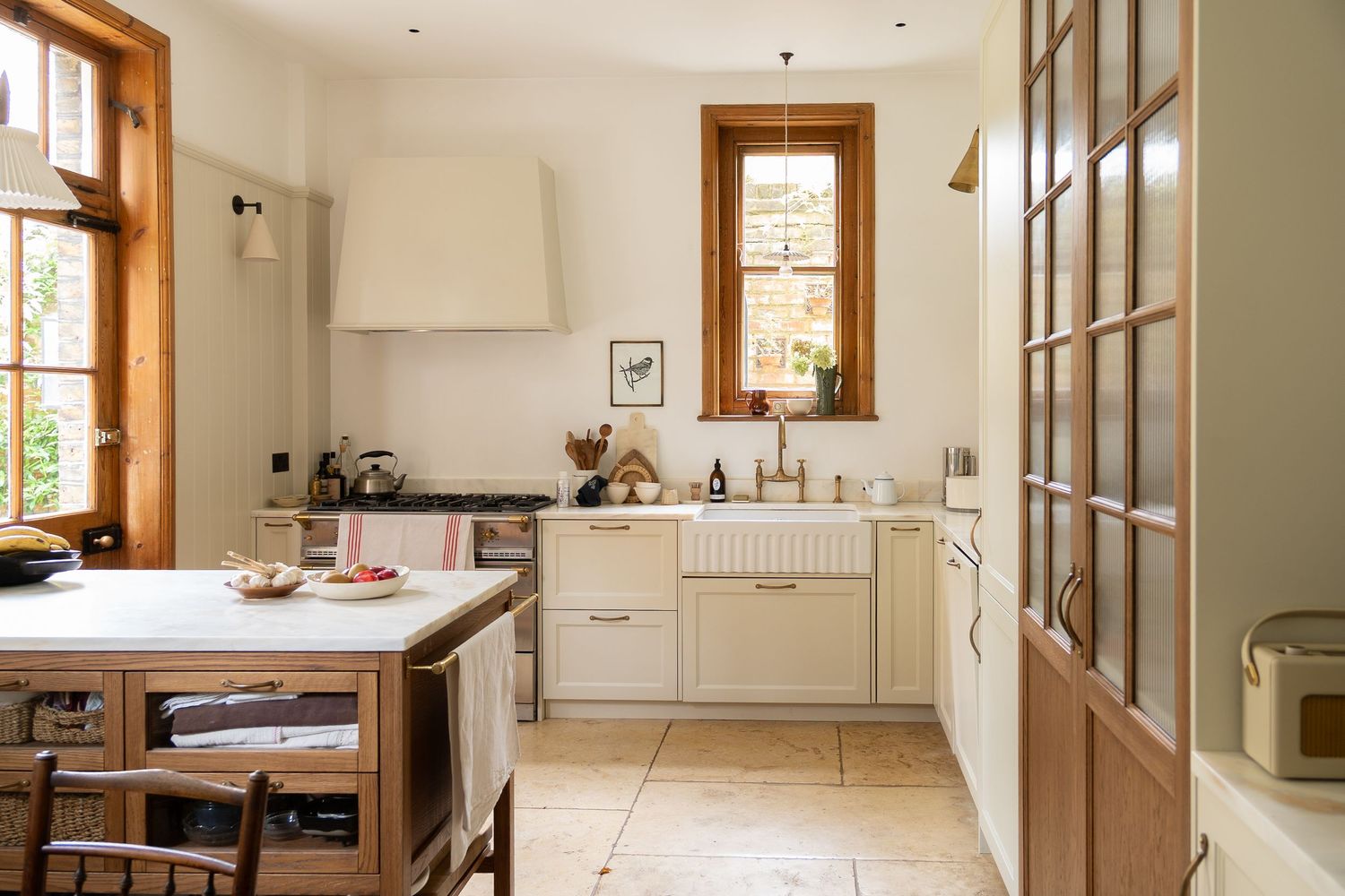 Cozy cream kitchen featuring a farmhouse sink, wood-trimmed windows, limestone floors, and central island.