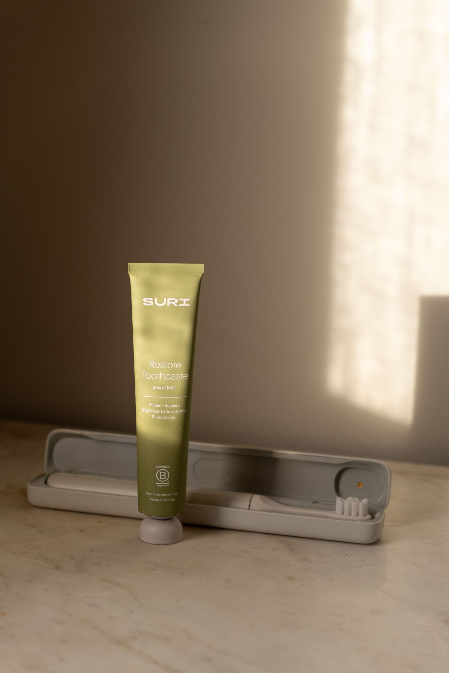SURI Restore toothpaste tube in olive green packaging beside a grey toothbrush travel case.
