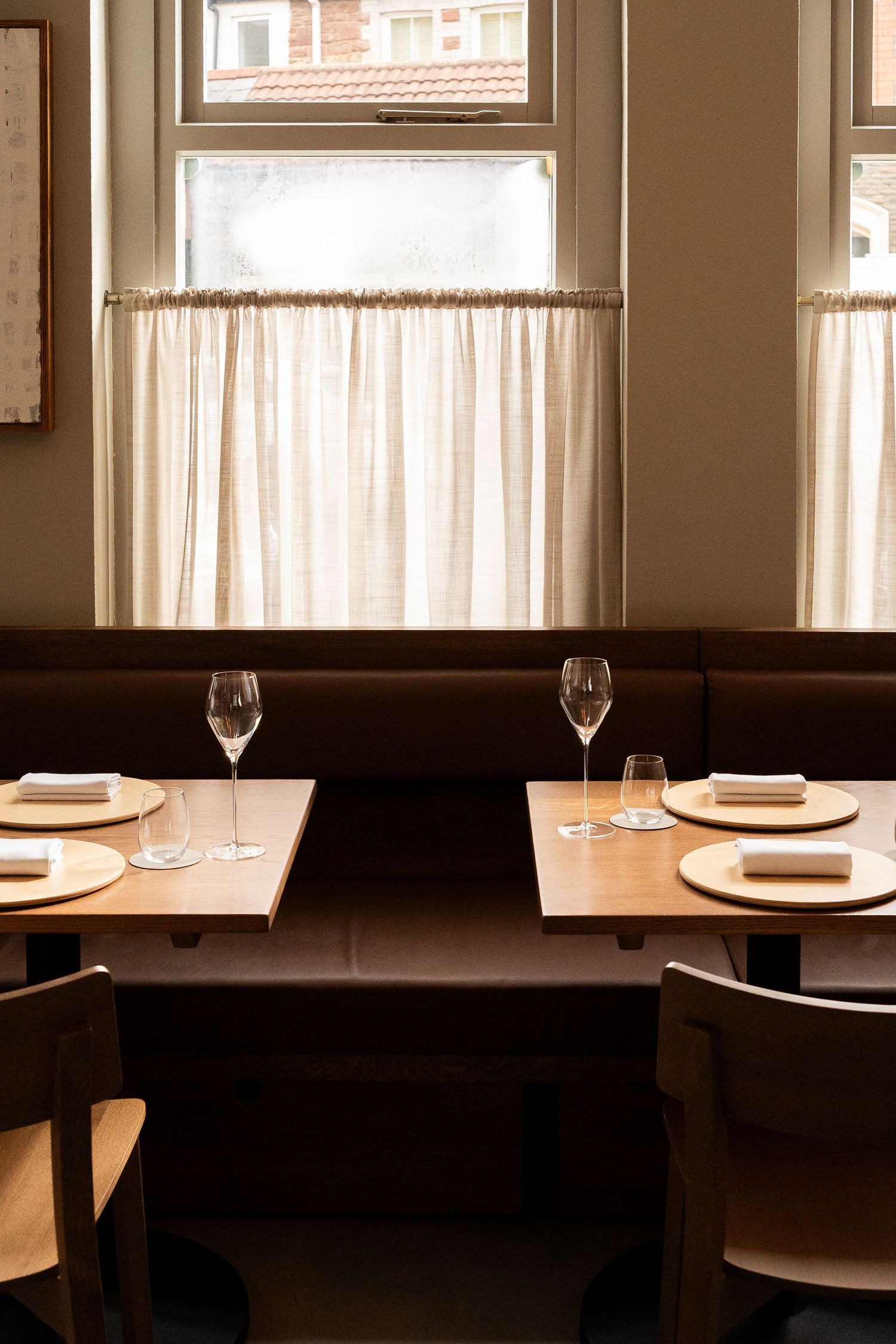 Elegant restaurant booth seating with linen curtains, set tables with wine glasses and white napkins by a window.