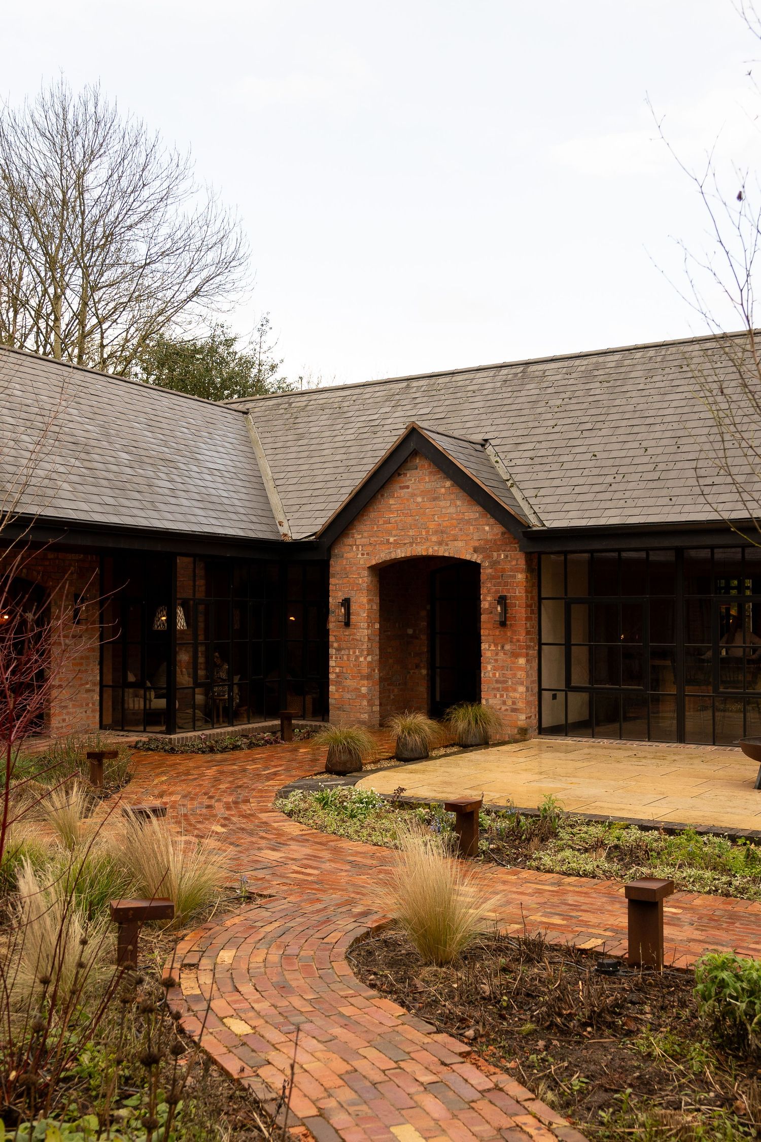Red brick country house with arched entrance, steel-framed windows, slate roof, and curved brick pathway through landscaped garden.