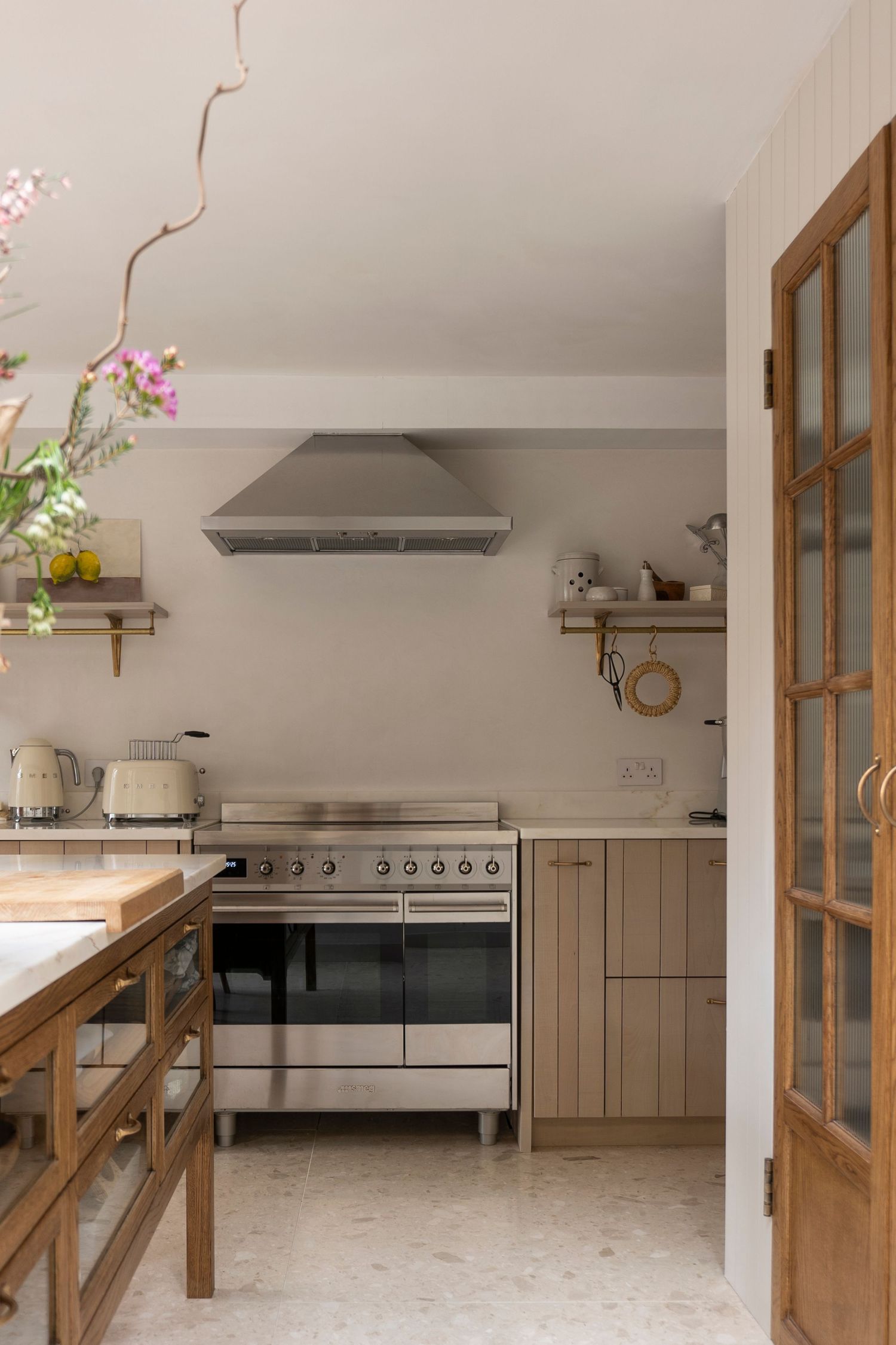 Neutral kitchen with stainless steel range, wood cabinetry, open shelves, terrazzo floor, and glass-paneled door.