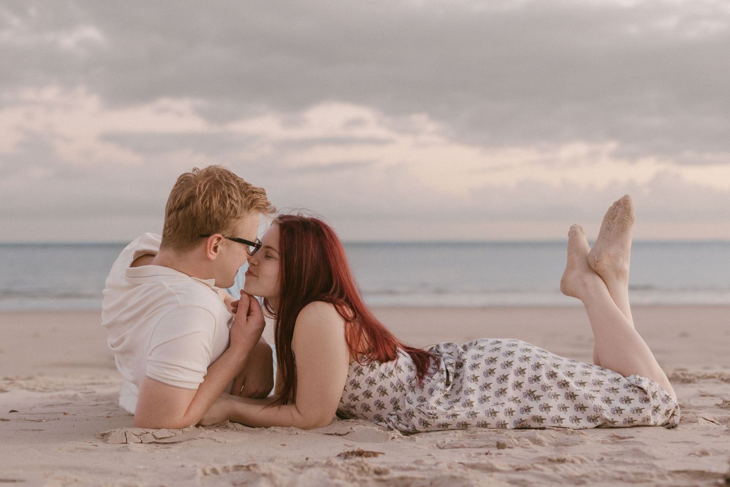 A couple lies on sandy beach facing each other in a tender romantic moment.