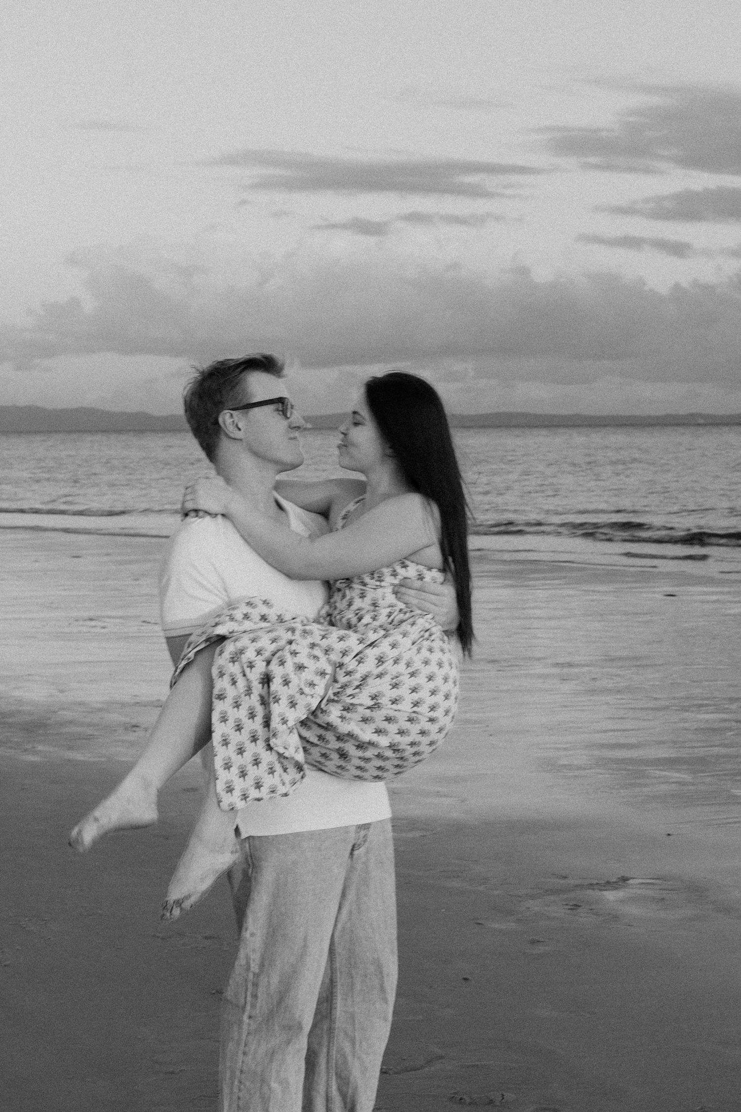 Man carries woman in his arms on a beach at sunset in black and white photo.