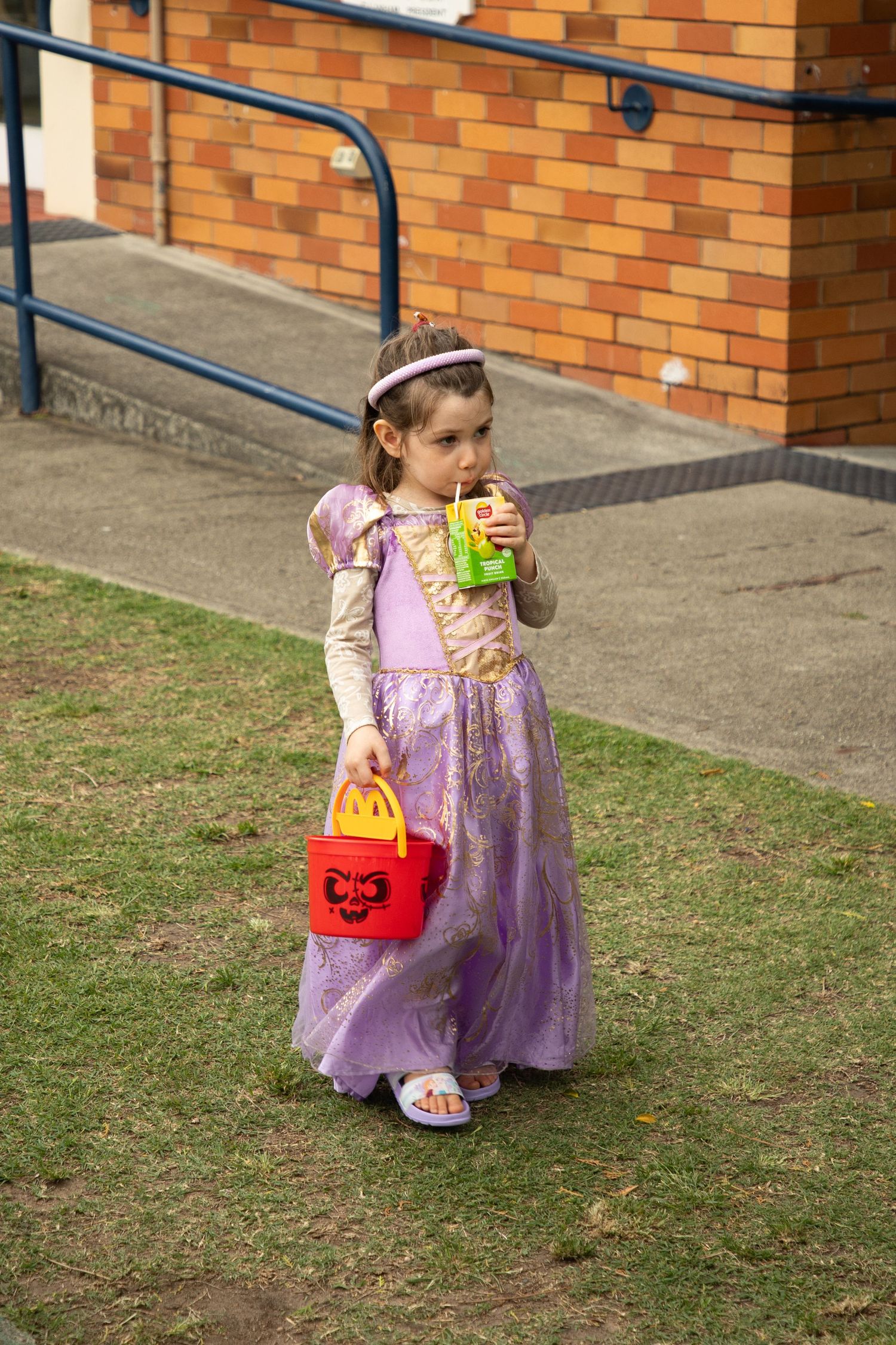 Young girl dressed as Rapunzel holds a Halloween bucket and drink at an outdoor event.