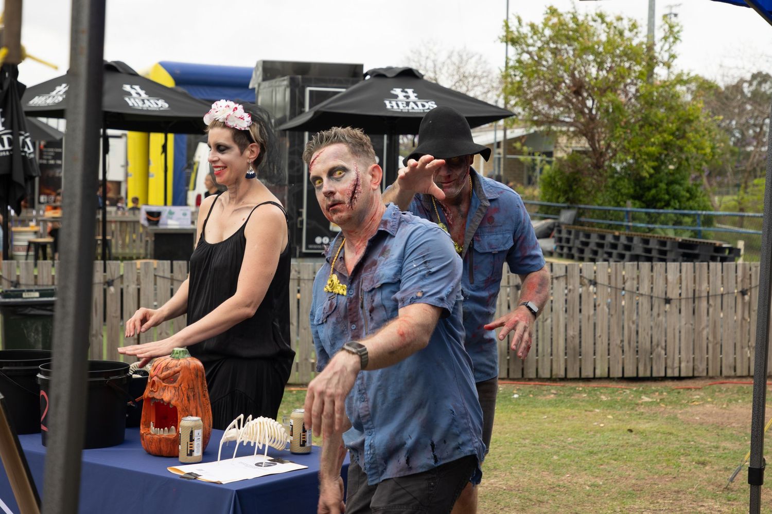 Three people in zombie Halloween makeup and costumes at outdoor festival booth.