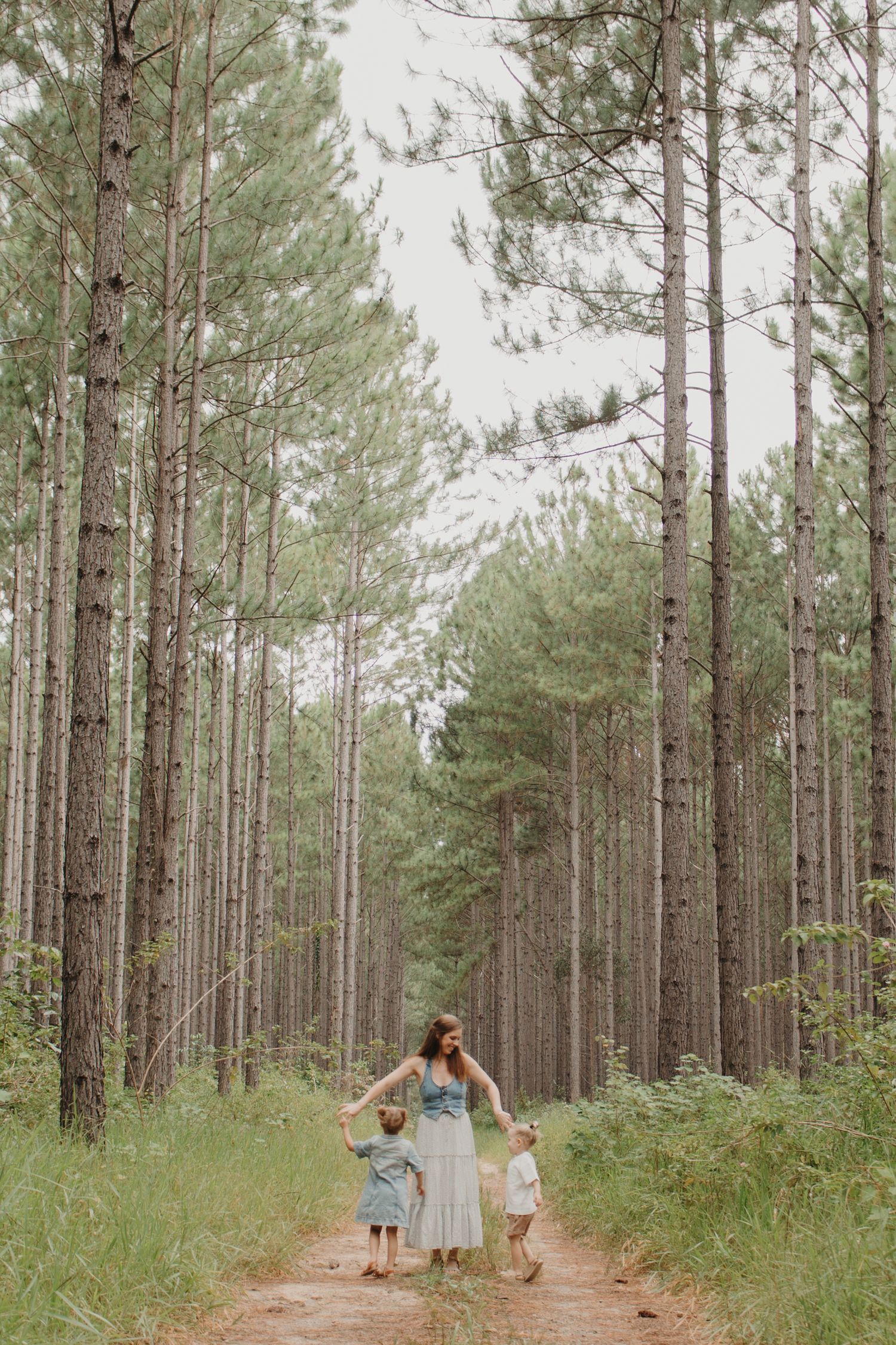 A mother holds hands with her two young children while walking away down a pine forest trail.