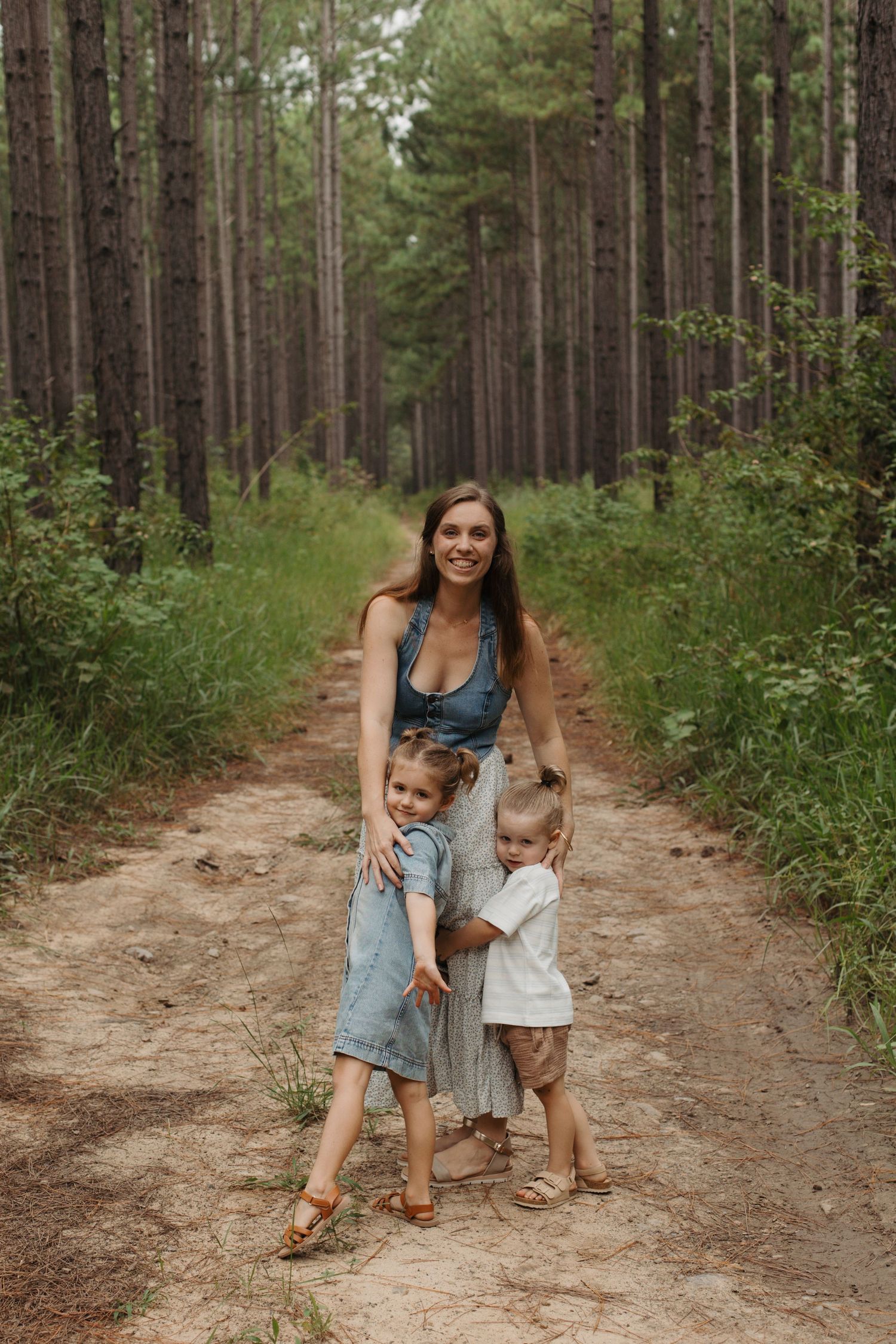 Smiling mother posing with two young children on a dirt path in a pine forest.