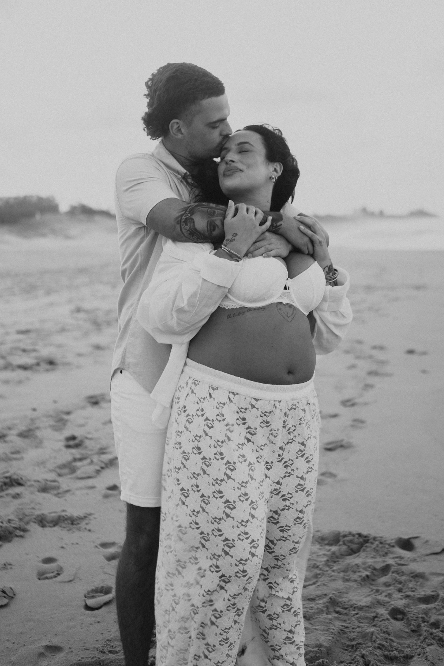 Black and white photo of a couple embracing on a beach, woman with baby bump in lace skirt, man kissing her cheek.