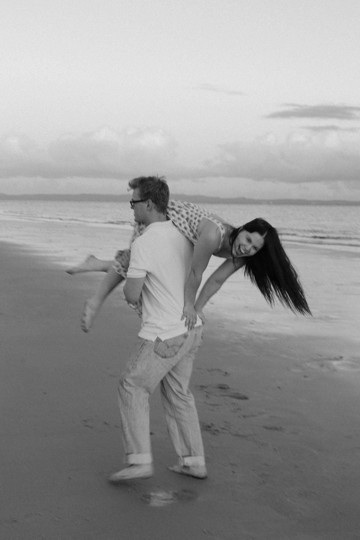 A man playfully carries a laughing woman over his shoulder on a beach at dusk.