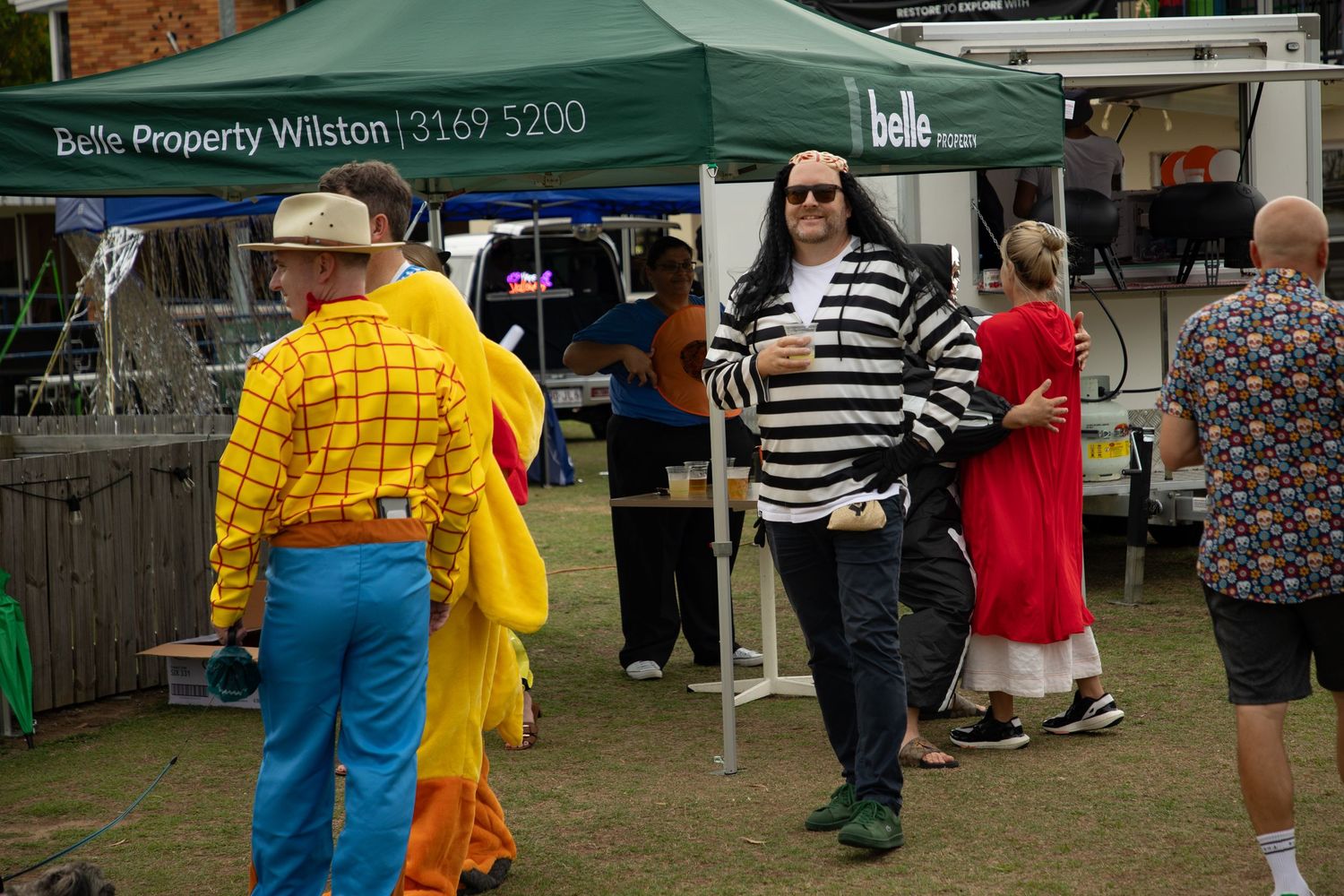 Adults in Halloween costumes including Woody and a prisoner socializing near a Belle Property Wilston tent.