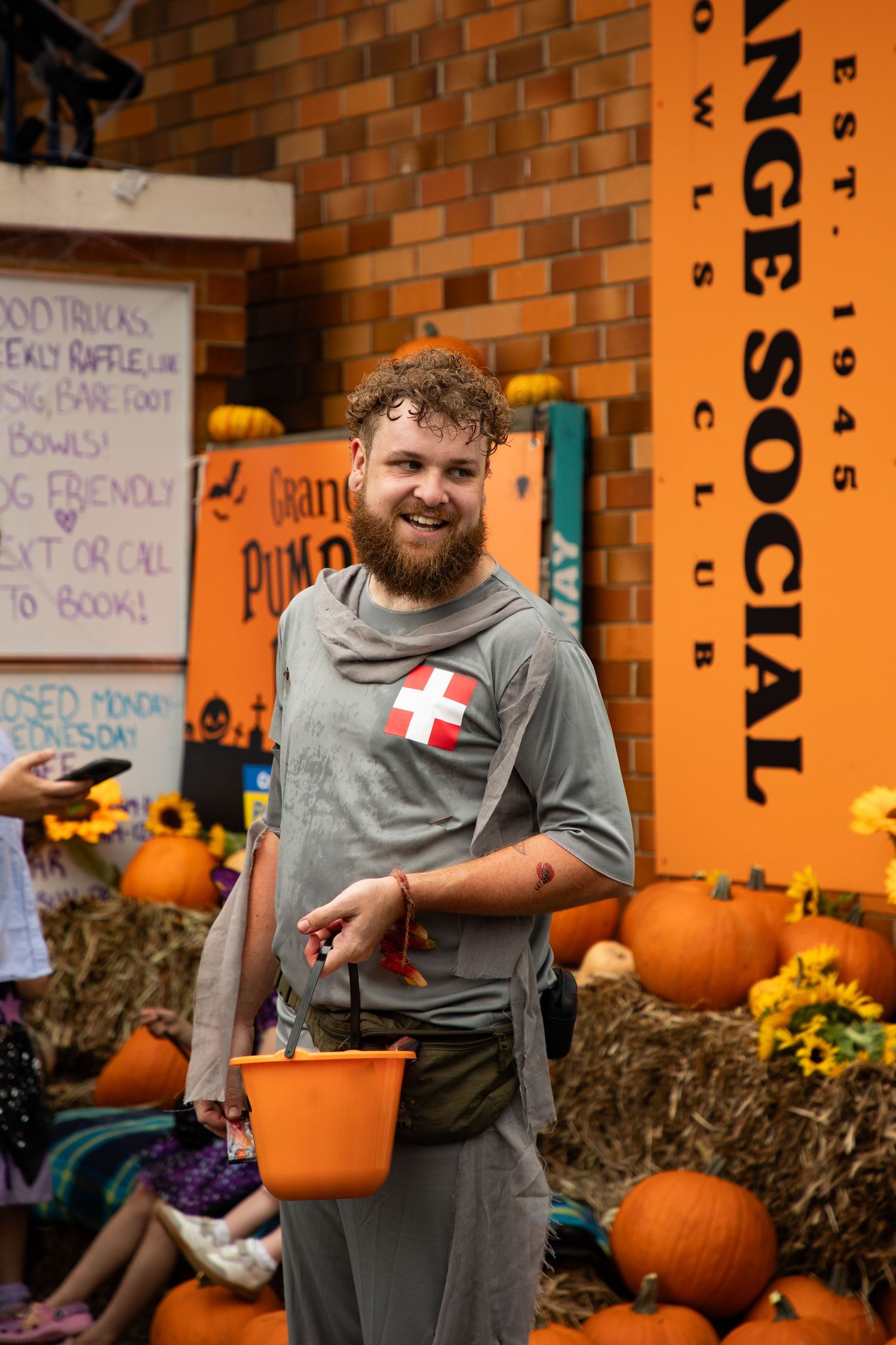 Bearded man in Halloween costume smiles holding orange bucket surrounded by pumpkins at festival.