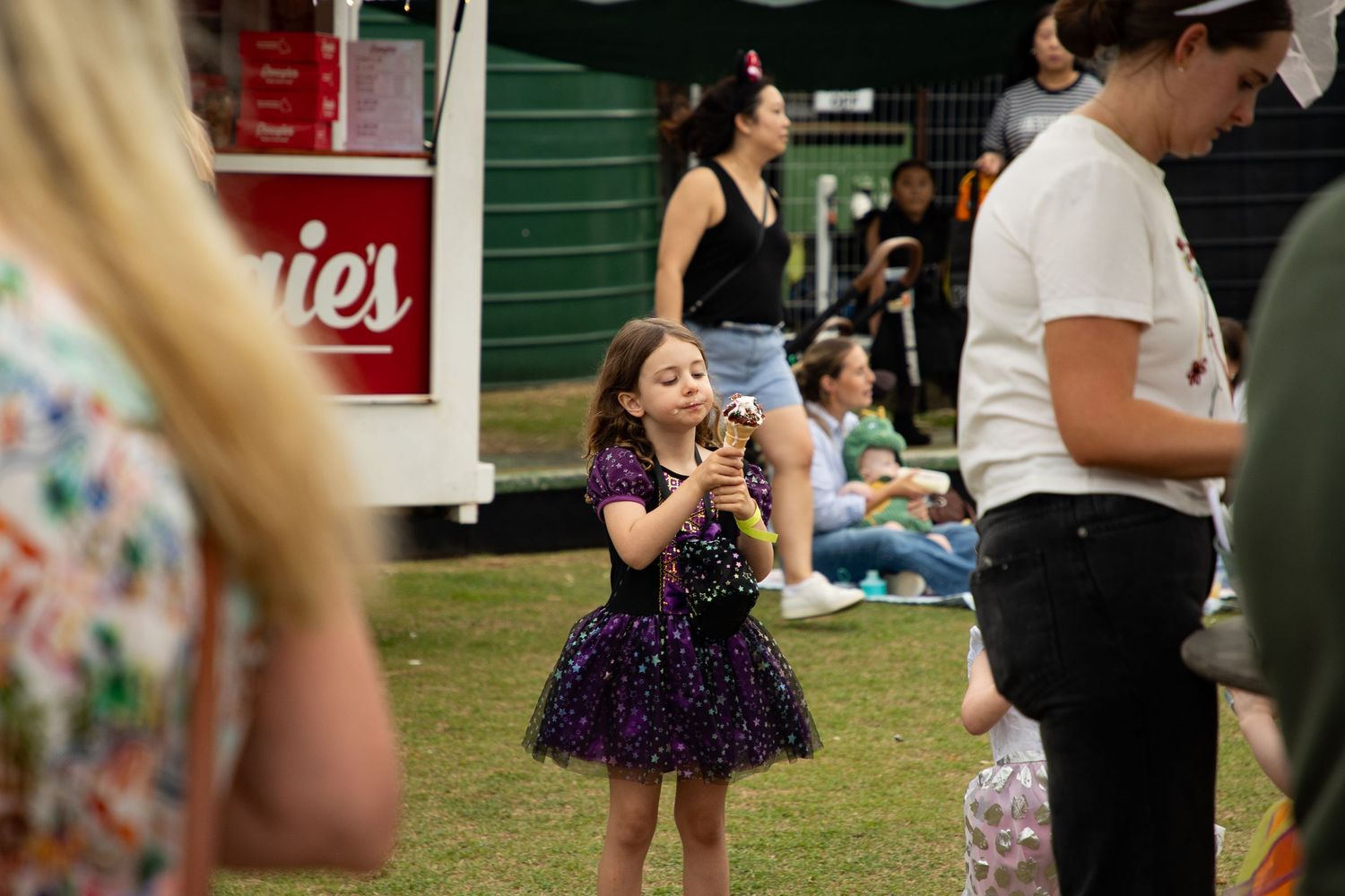 A young girl in a purple sparkly dress enjoys a treat at a busy outdoor festival event.