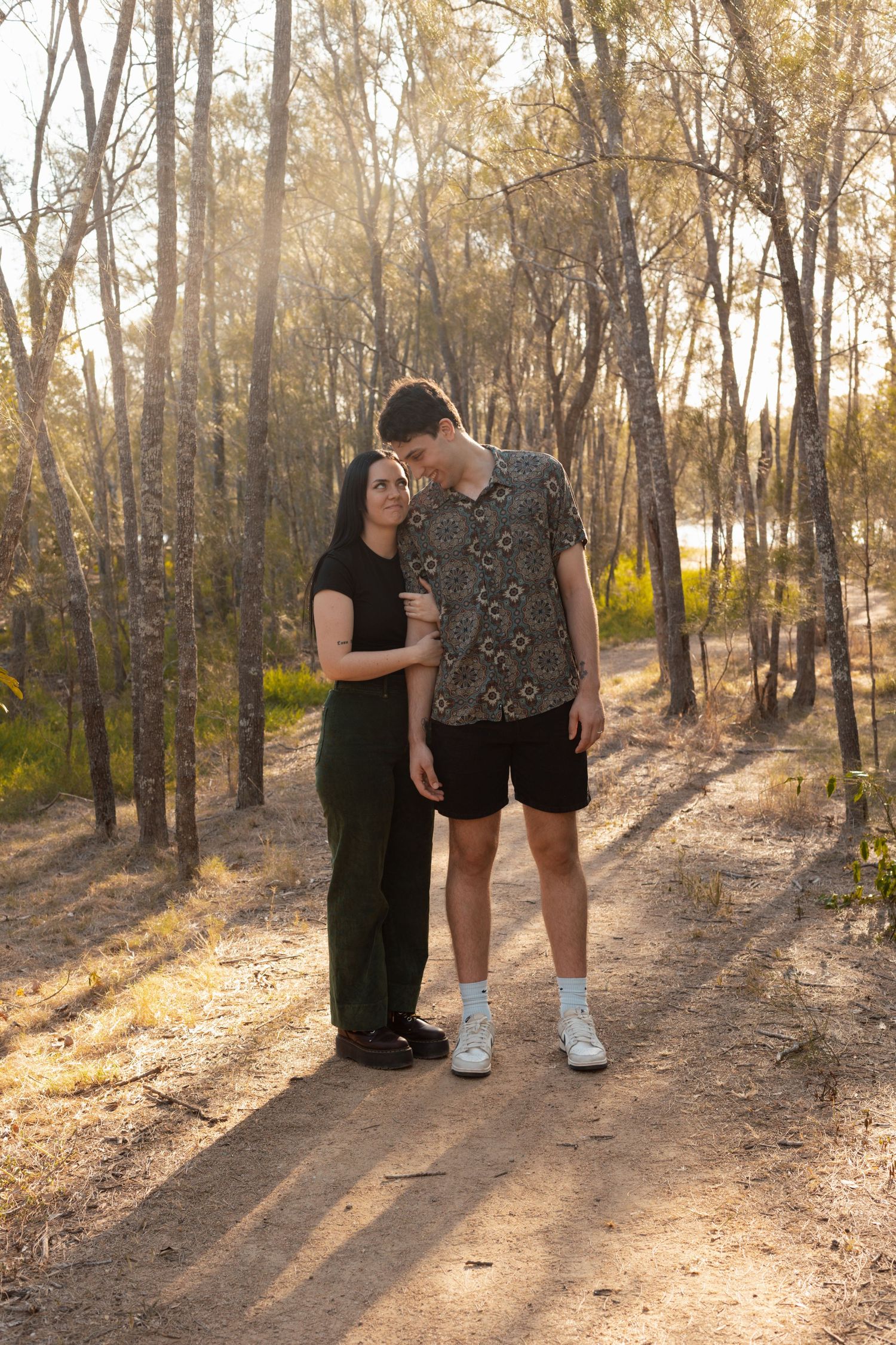 Young couple standing close together on a forest path bathed in warm golden sunlight.