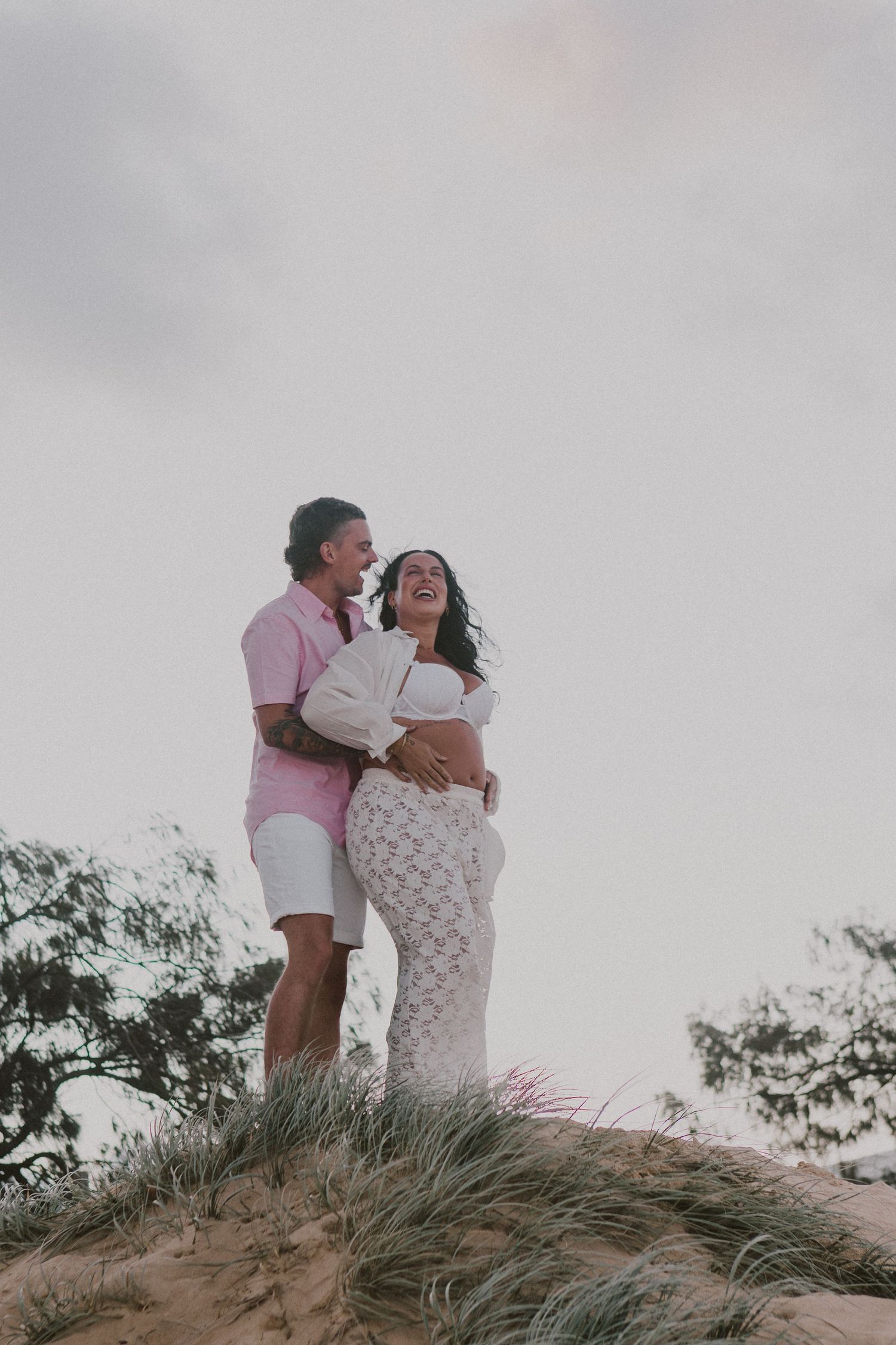 Couple embracing on sandy dune at sunset, pregnant woman in white lace dress with partner in pink shirt.