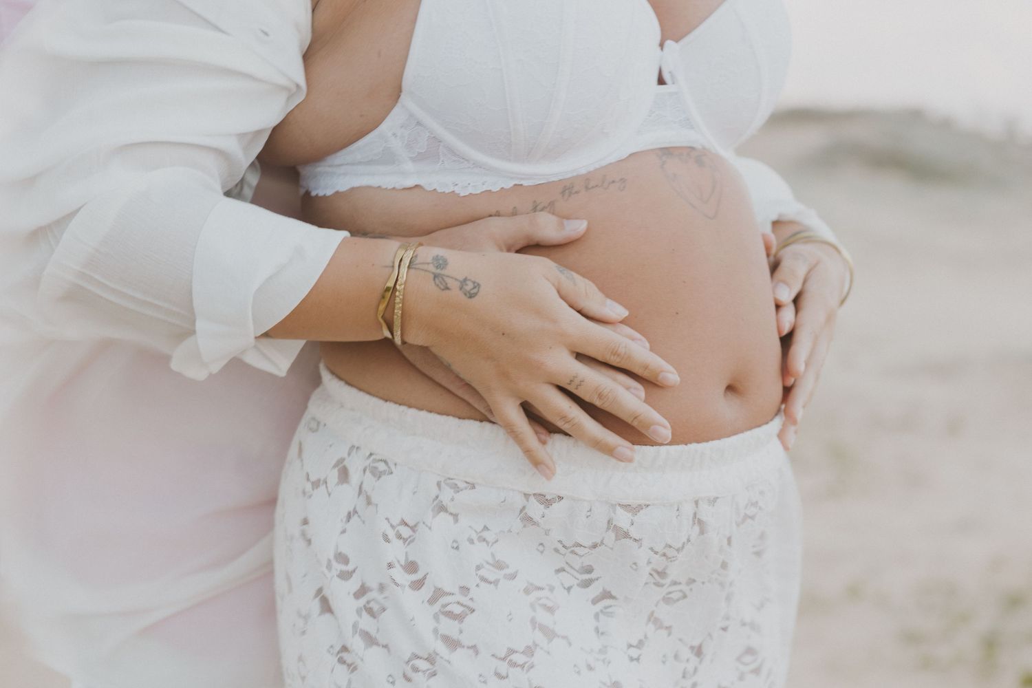 Couple tenderly cradling pregnant belly outdoors, woman in white lace skirt and bra top during maternity photoshoot.