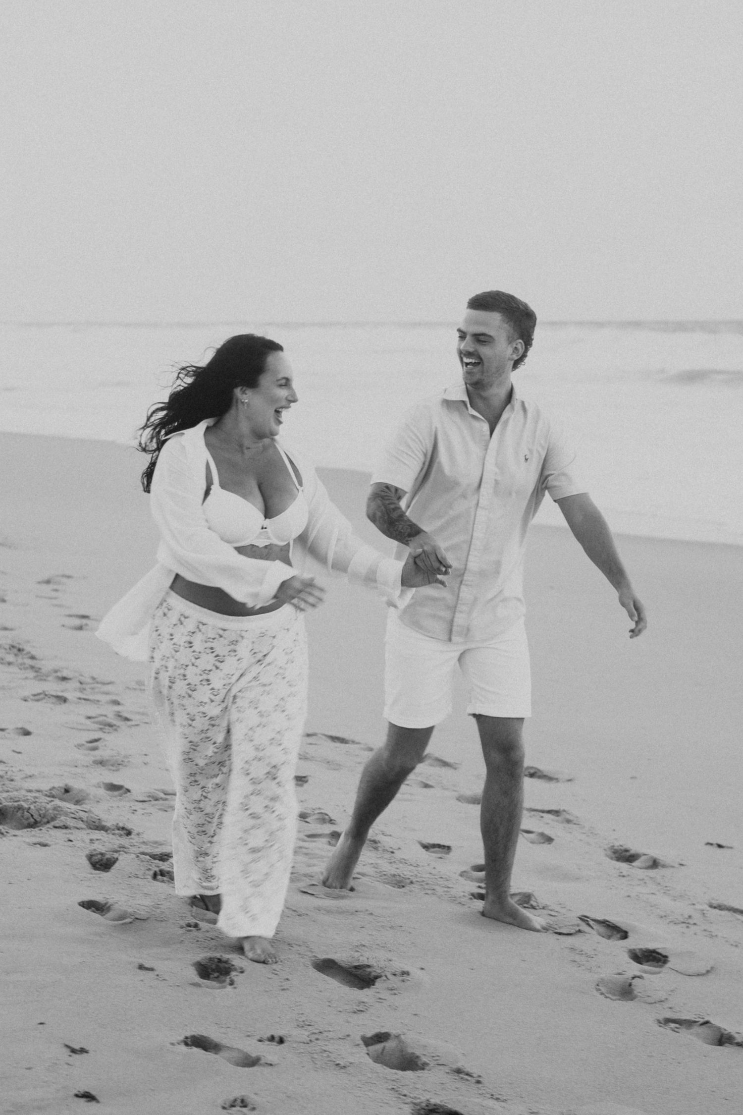 Couple laughing and holding hands while walking barefoot on sandy beach in matching white outfits, black and white photo.