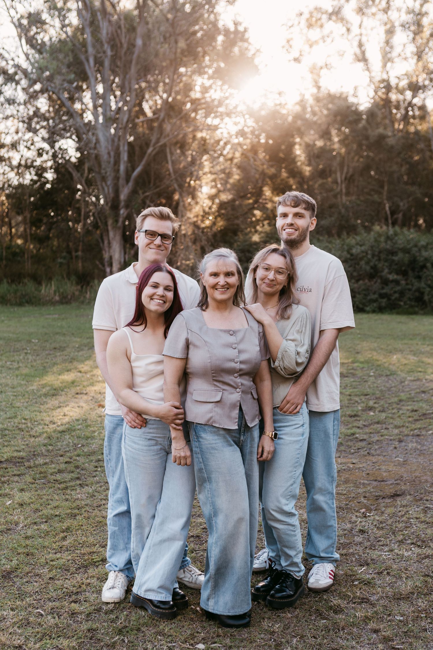 Family of five smiling together outdoors at golden hour, wearing casual jeans and neutral tones in a lush green park.