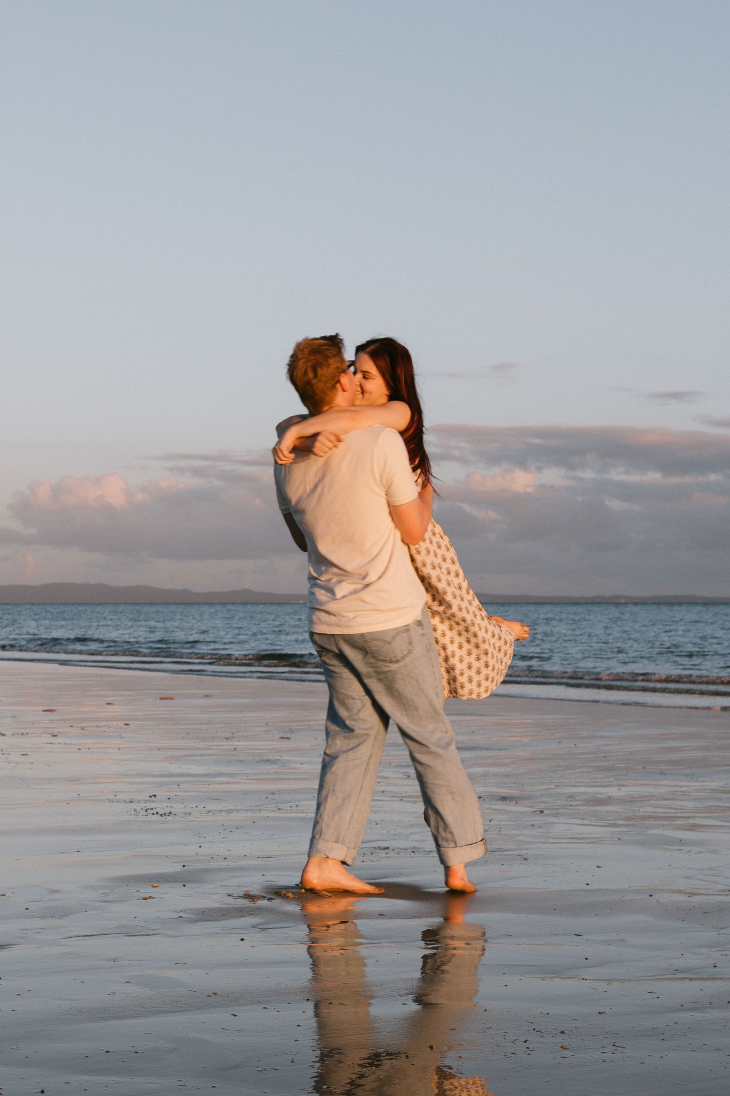Couple sharing a romantic kiss while embracing on a reflective beach at golden hour sunset.