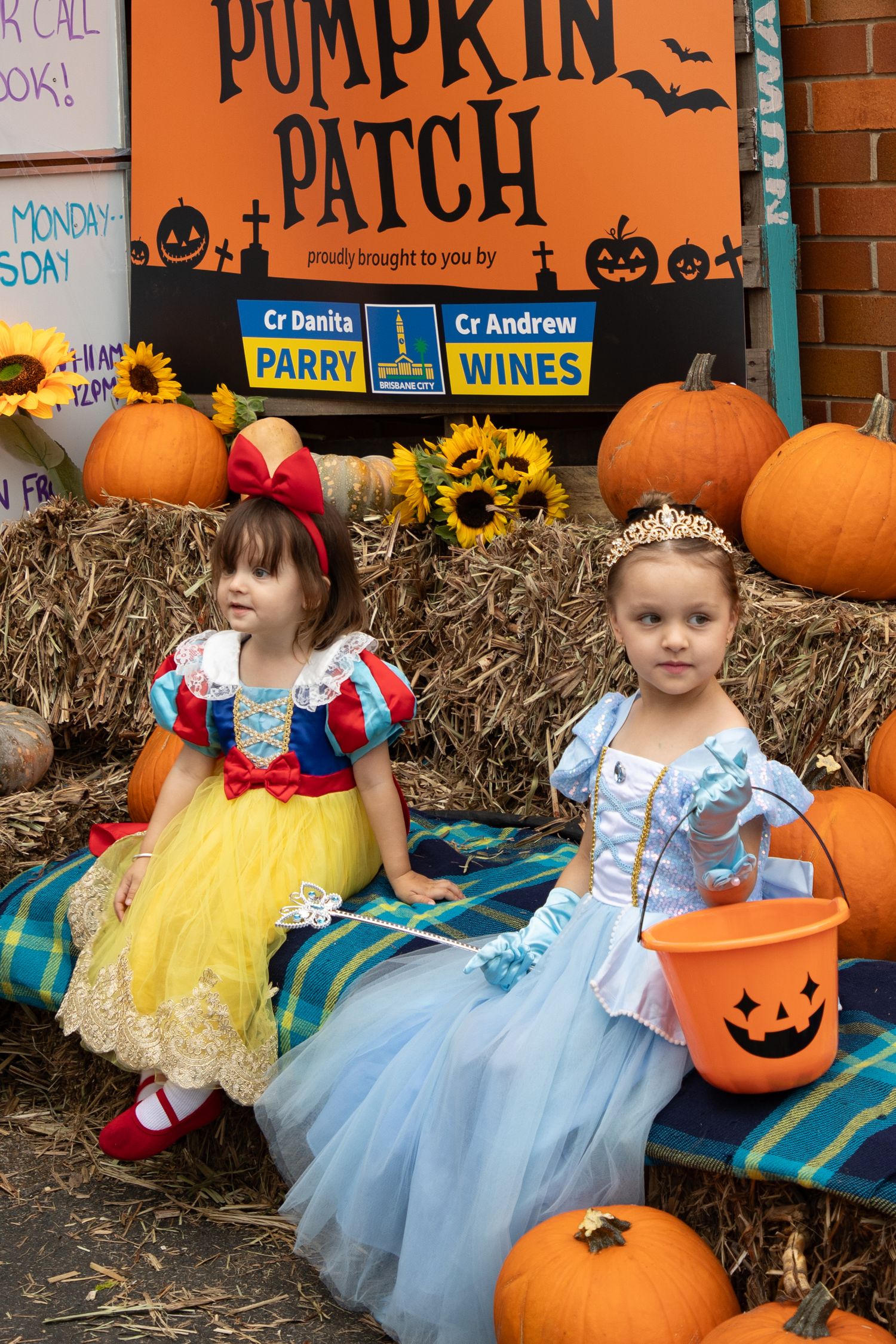 Two young girls dressed as Snow White and Cinderella sit at a Halloween pumpkin patch display.