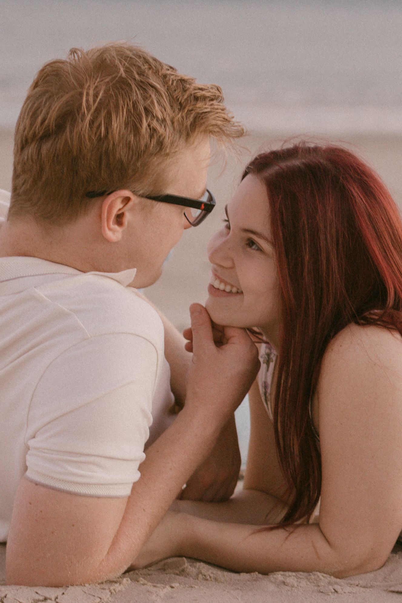 A young couple smiling at each other while lying on a sandy beach, sharing a romantic moment.