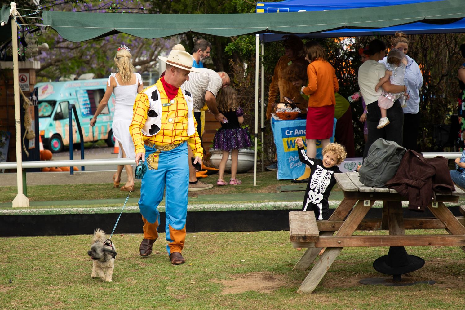 Man in Woody cowboy costume walks dog at Halloween festival while child in skeleton suit sits nearby.