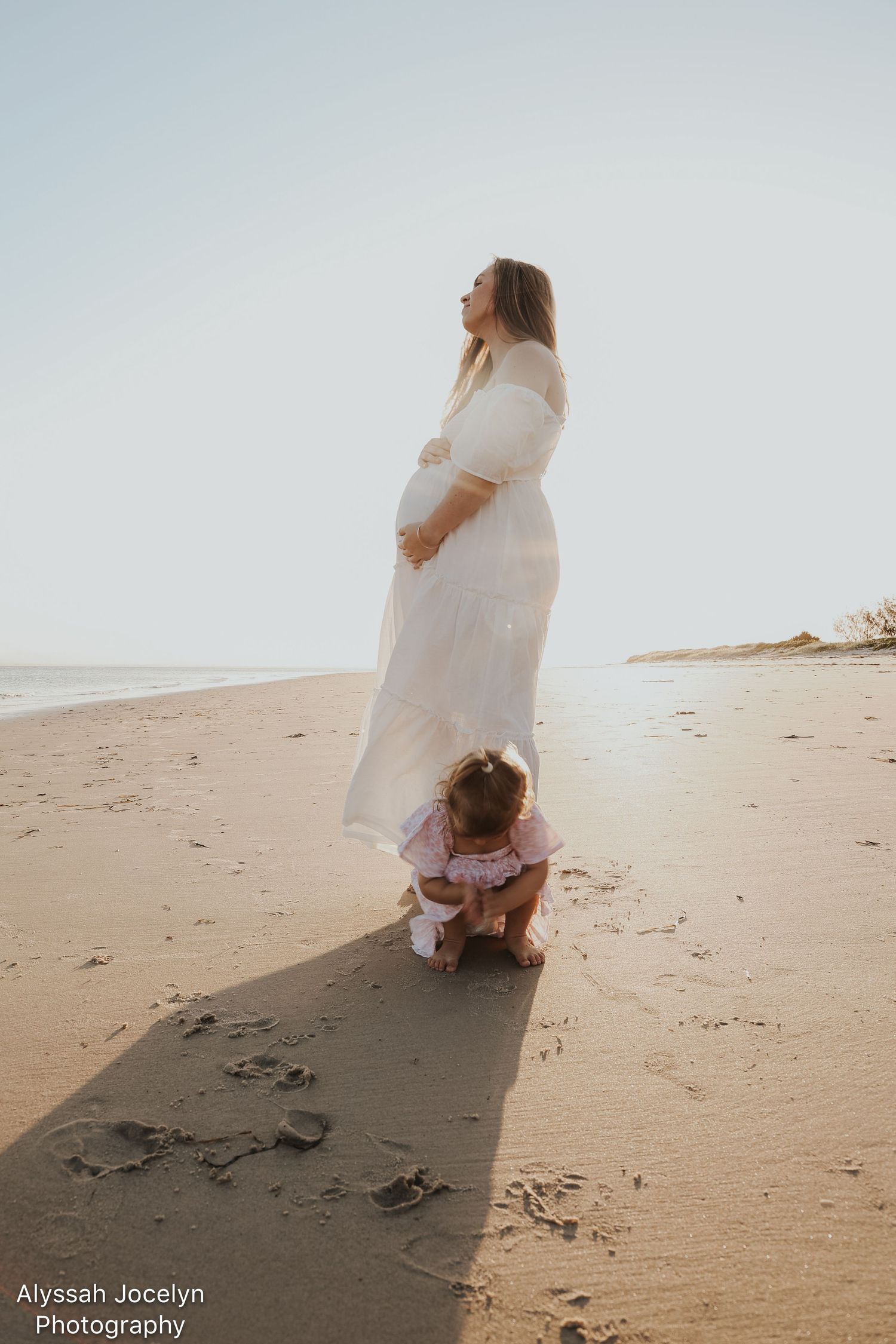 Pregnant woman in white dress on beach with toddler crouching in sand at sunset.