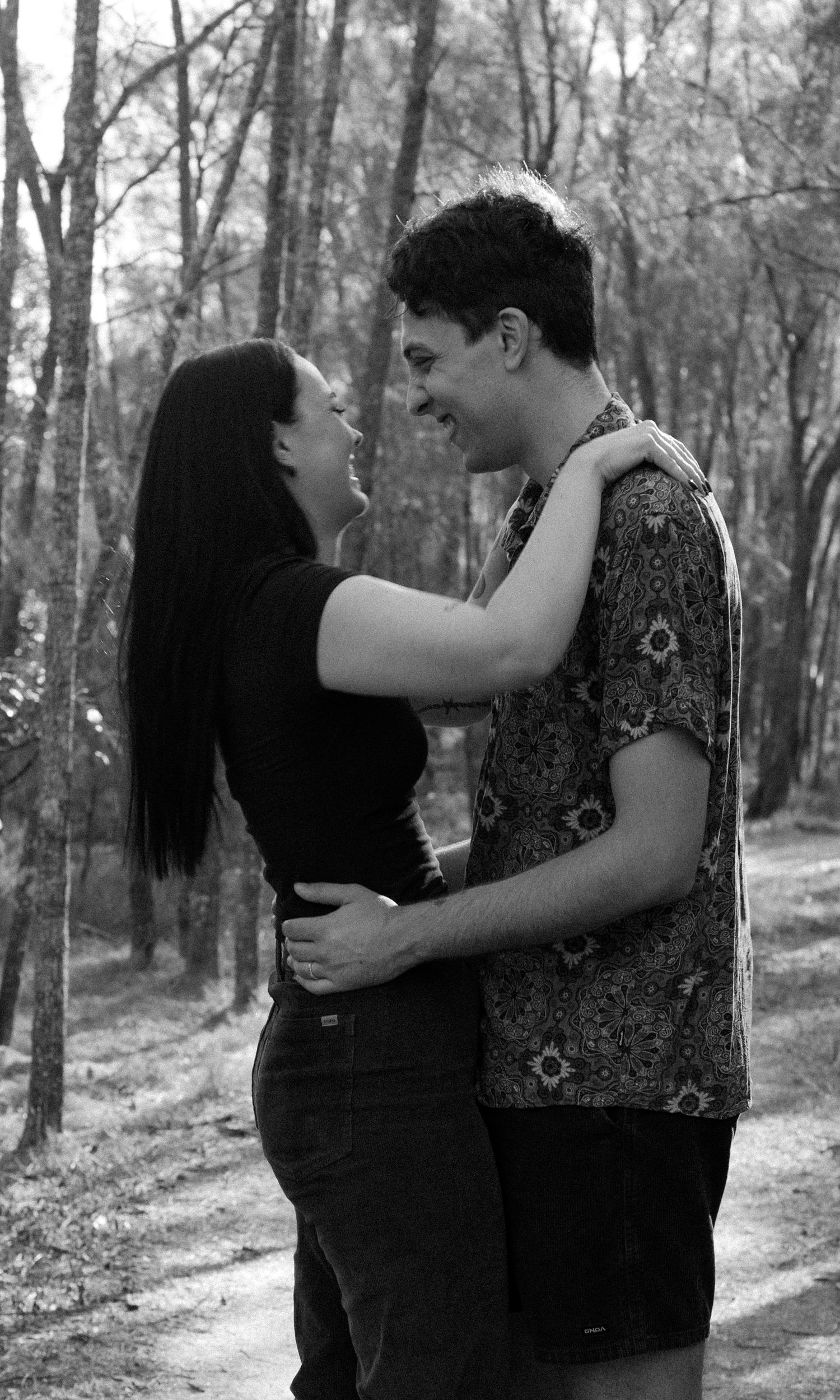Black and white photo of a young couple embracing and smiling at each other on a forest path.