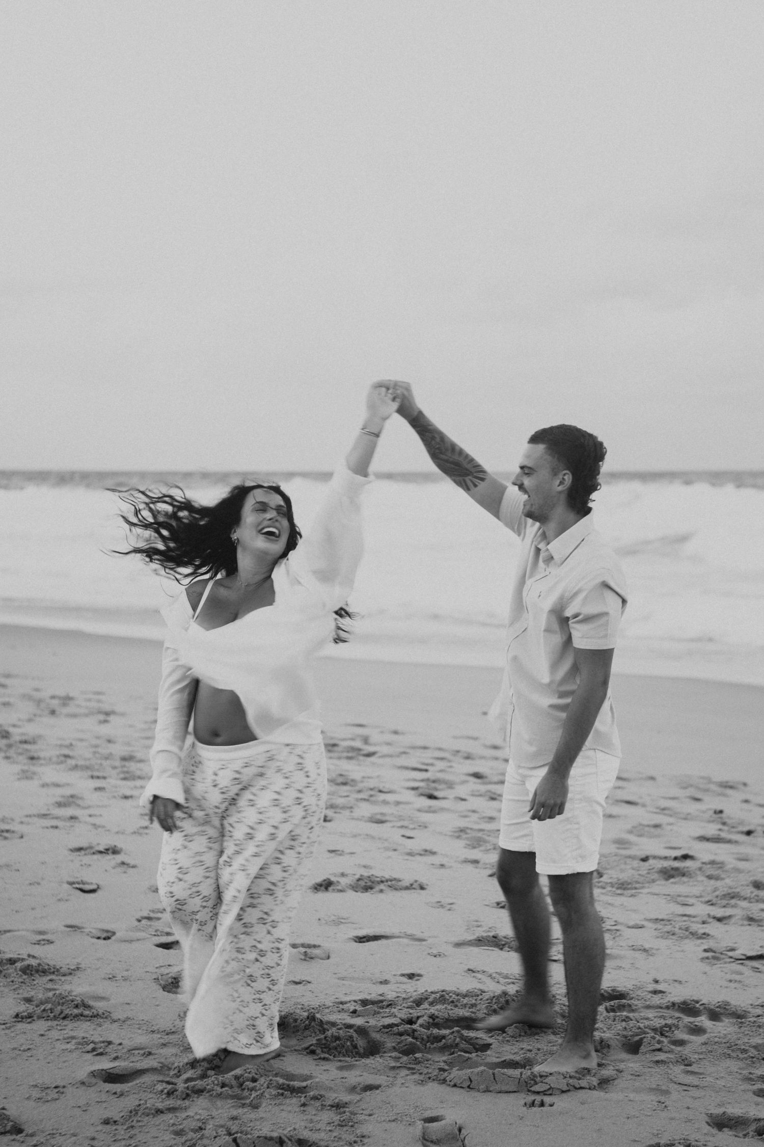 Black and white photo of a joyful couple dancing on the beach, woman laughing as man spins her hand.